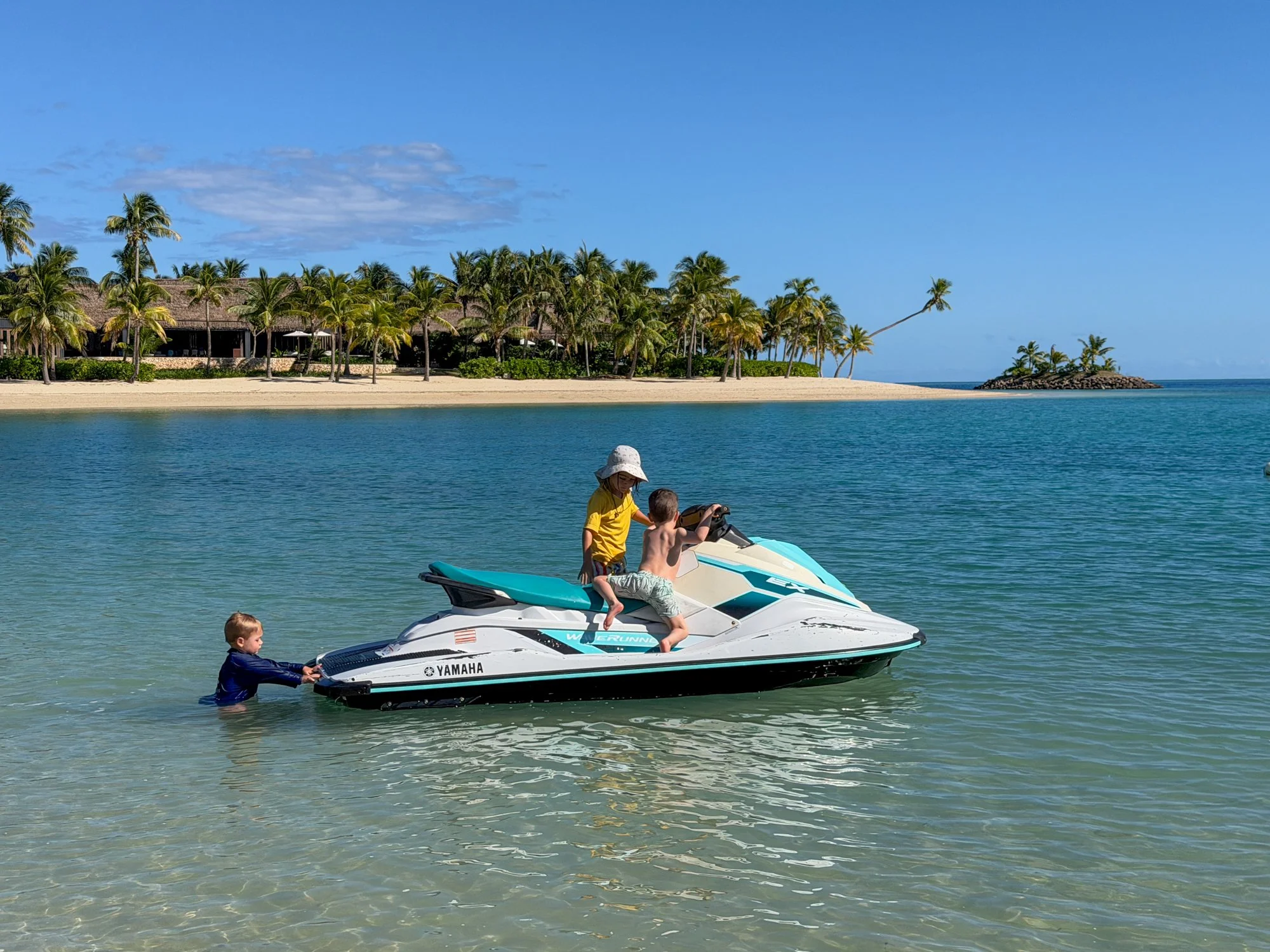 Young children play around a jet ski in the shallow turquoise lagoon of a Fiji resort.