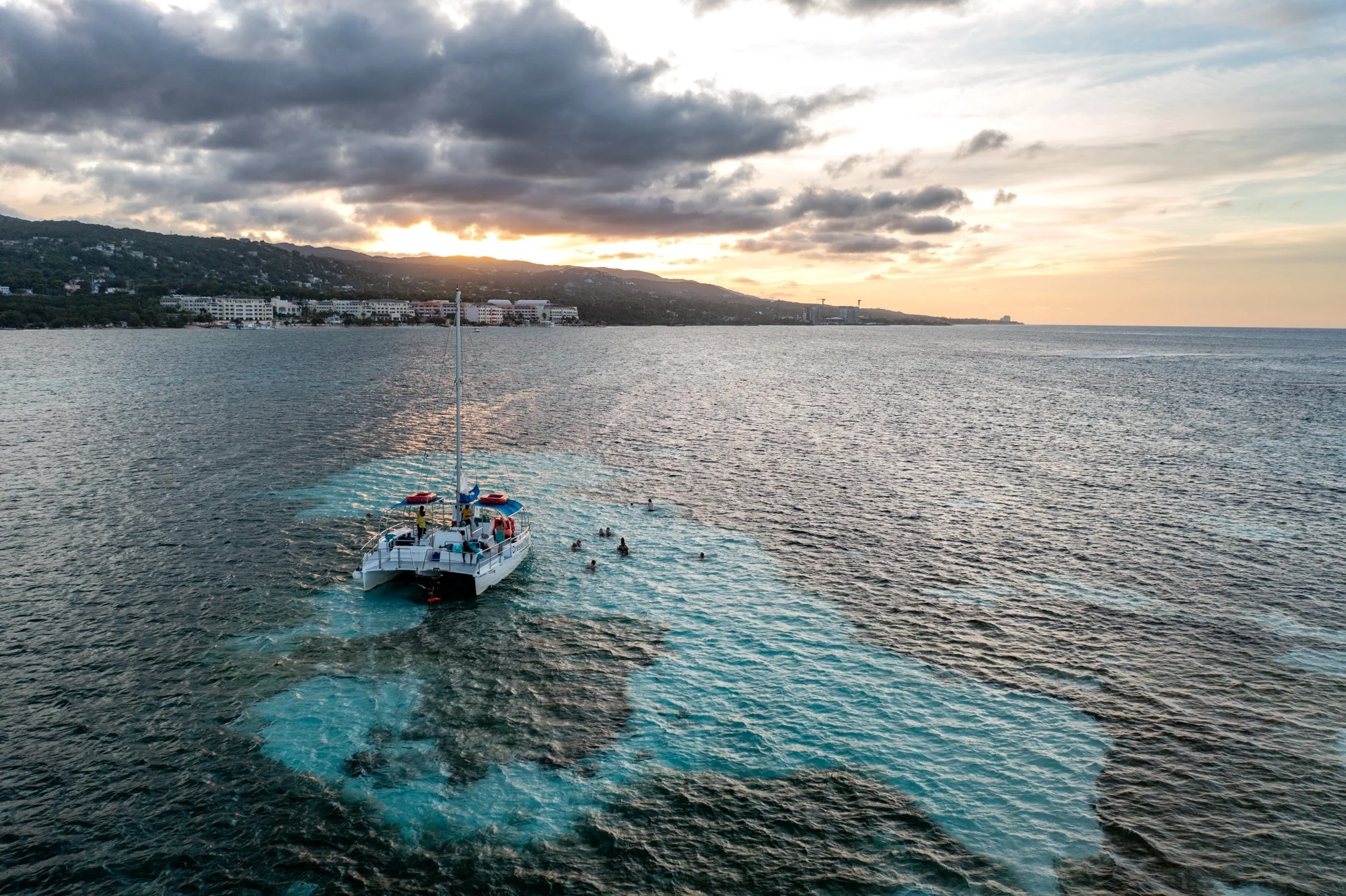 Aerial view of a catamaran and swimmers above a turquoise reef at sunset in Jamaica.