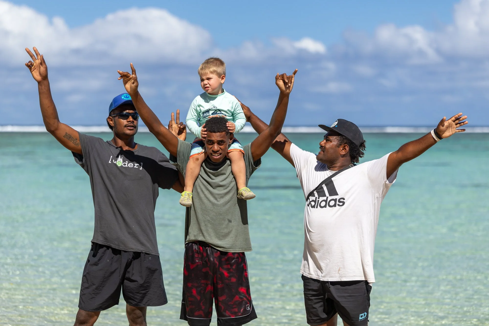 Three Fijian hosts celebrate on a tropical beach with a young child riding on one man’s shoulders.