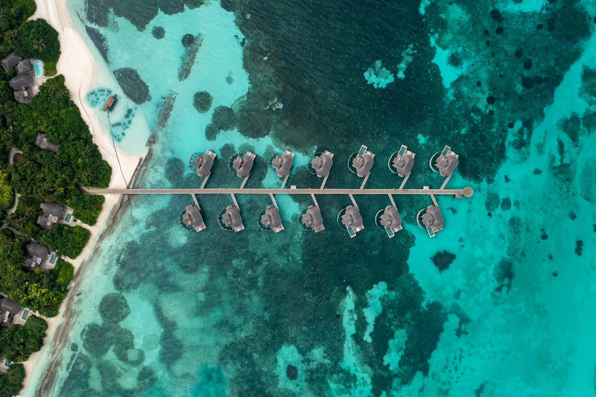 Top-down aerial view of overwater villas and a wooden jetty above a turquoise Maldives lagoon.