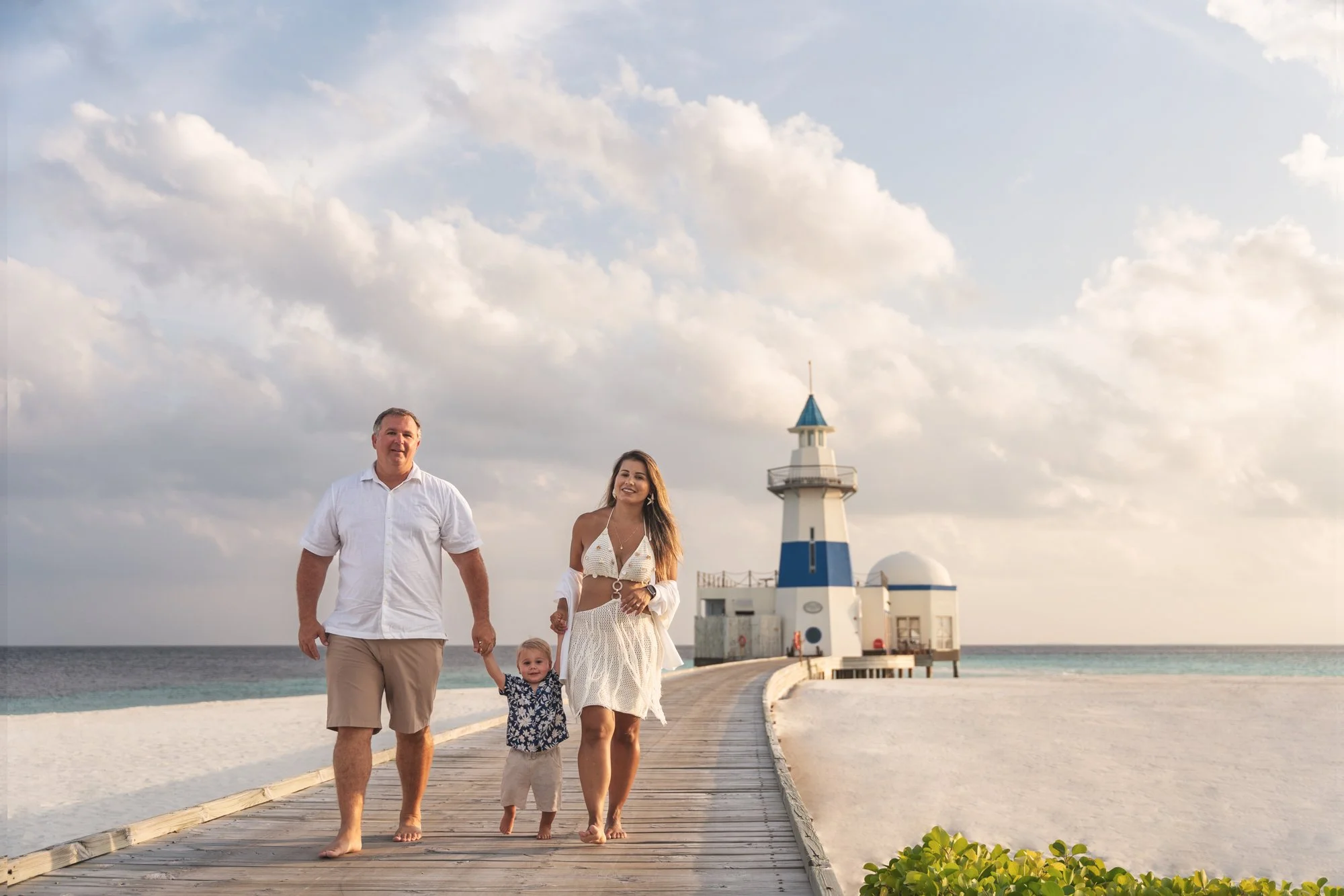 Family walking along a wooden boardwalk toward a blue-and-white Maldives lighthouse.