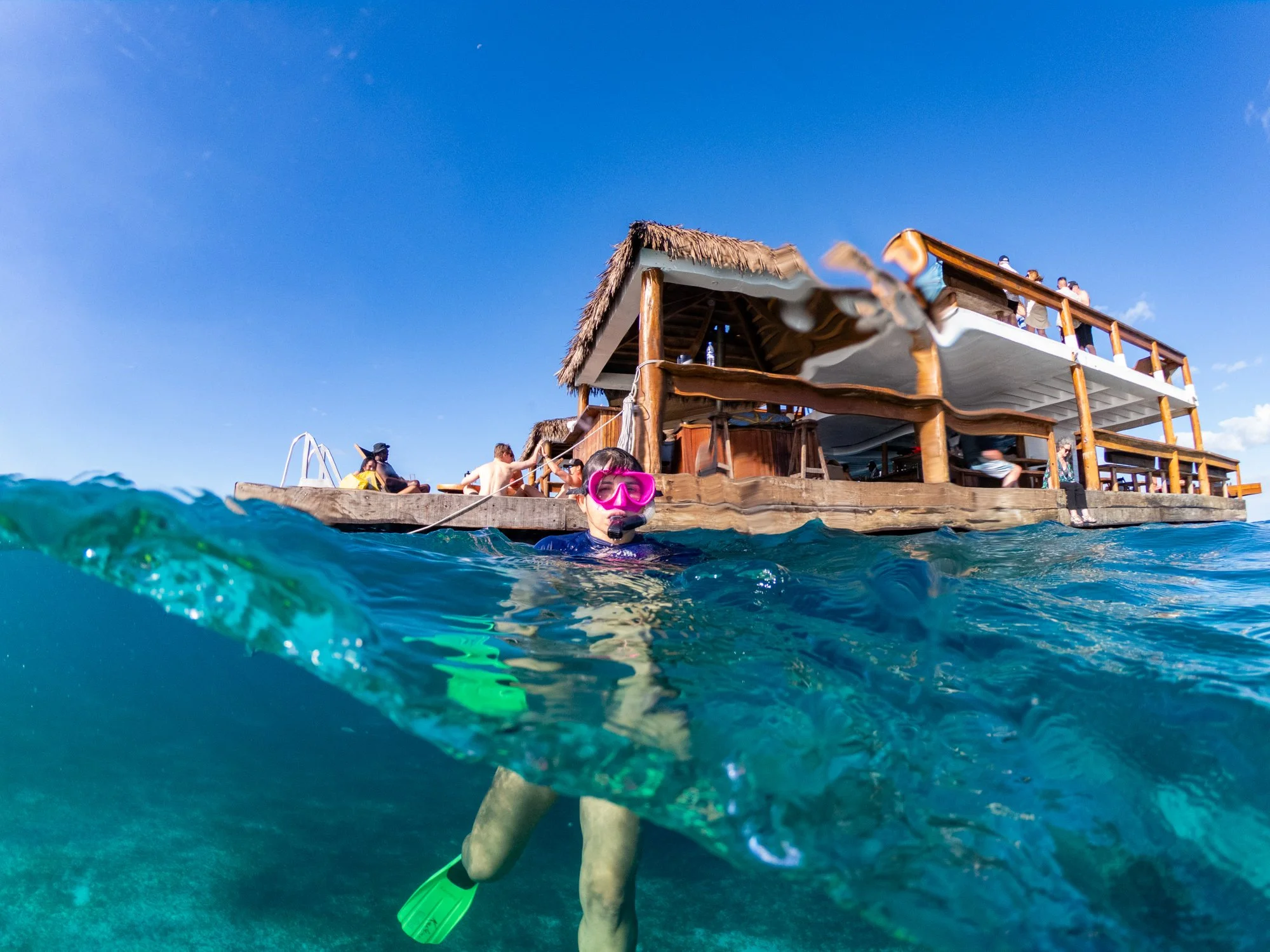 A snorkeler floats beside a thatched-roof restaurant anchored in clear Fijian water.