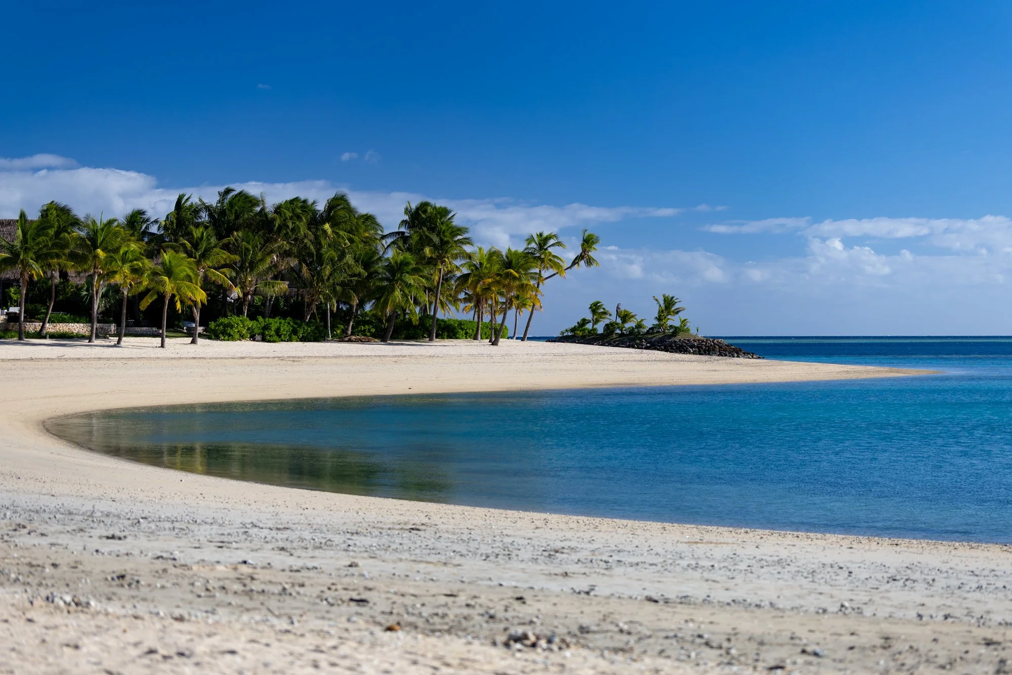 A quiet white-sand beach curves around a blue lagoon lined with palm trees in Fiji.
