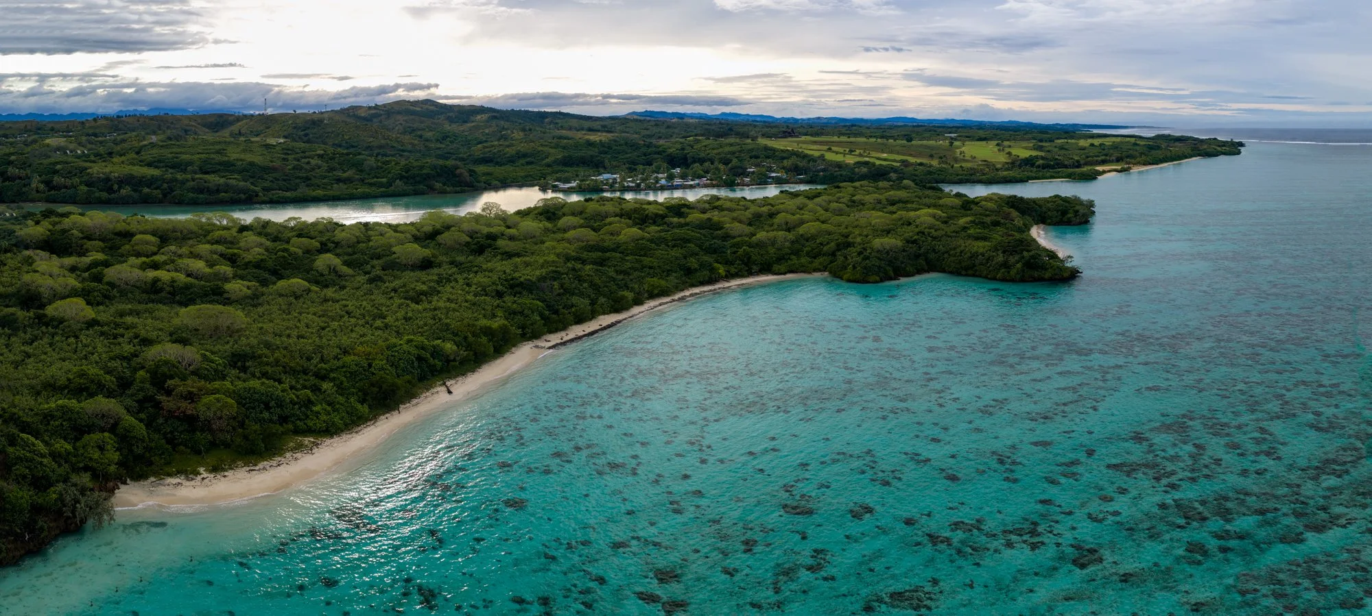 Panoramic aerial view of a forested Fijian coastline and clear turquoise coral lagoon.