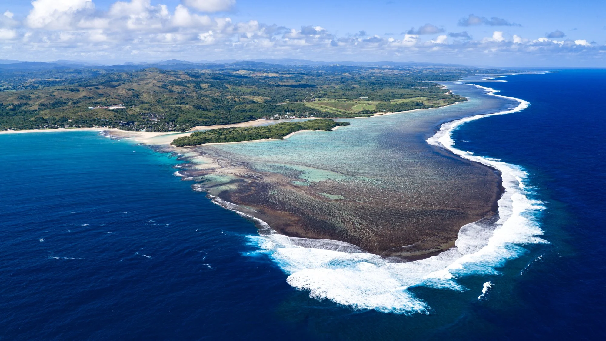 Aerial view of waves breaking along Fiji’s coral reef beside a lush green coastline.