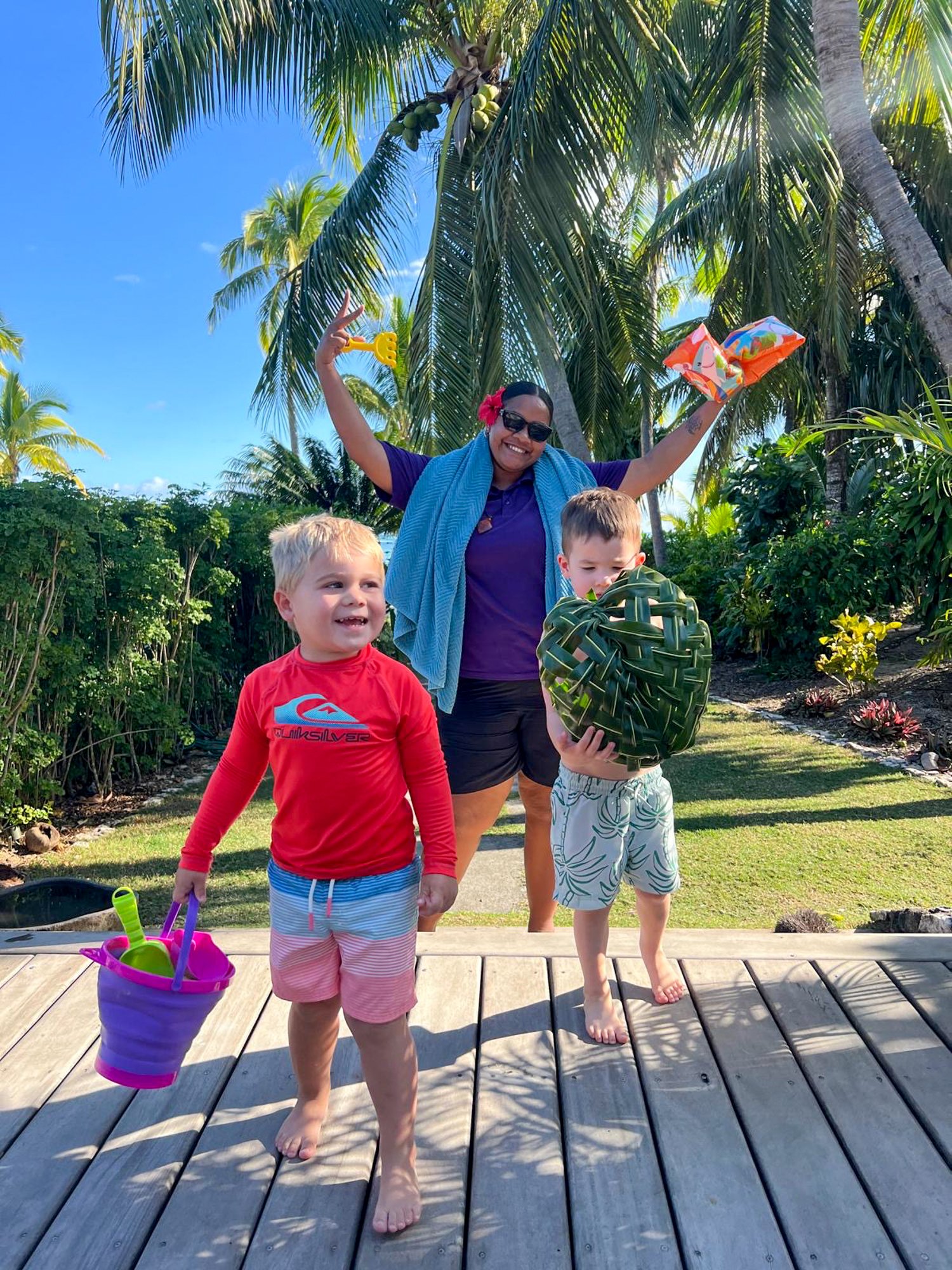 A smiling Fijian host welcomes two young children with a woven palm-leaf basket.