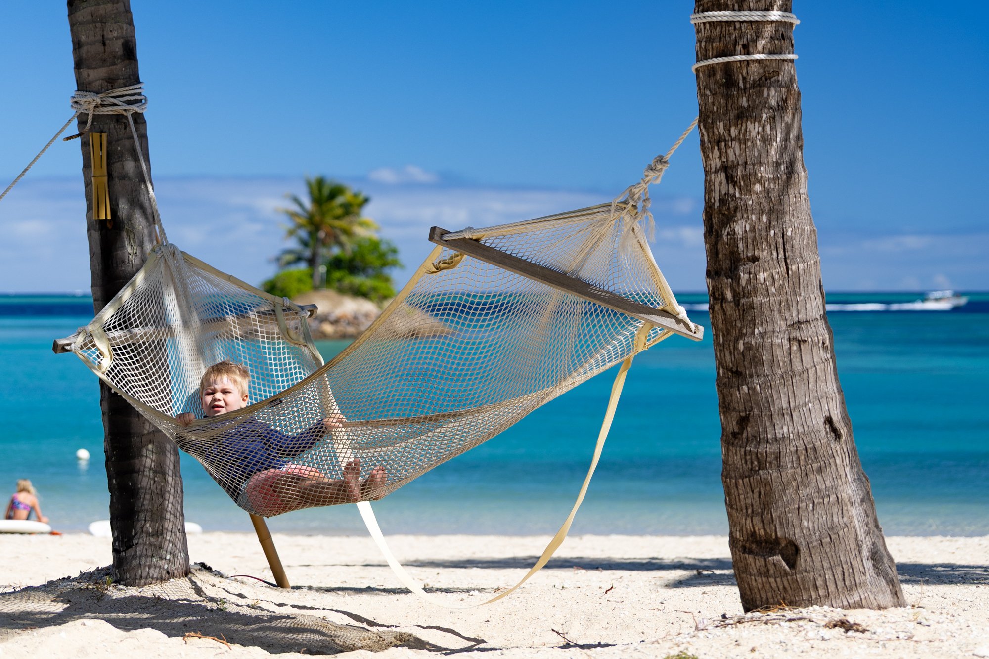 A young child relaxes in a hammock between palm trees overlooking the turquoise sea in Fiji.
