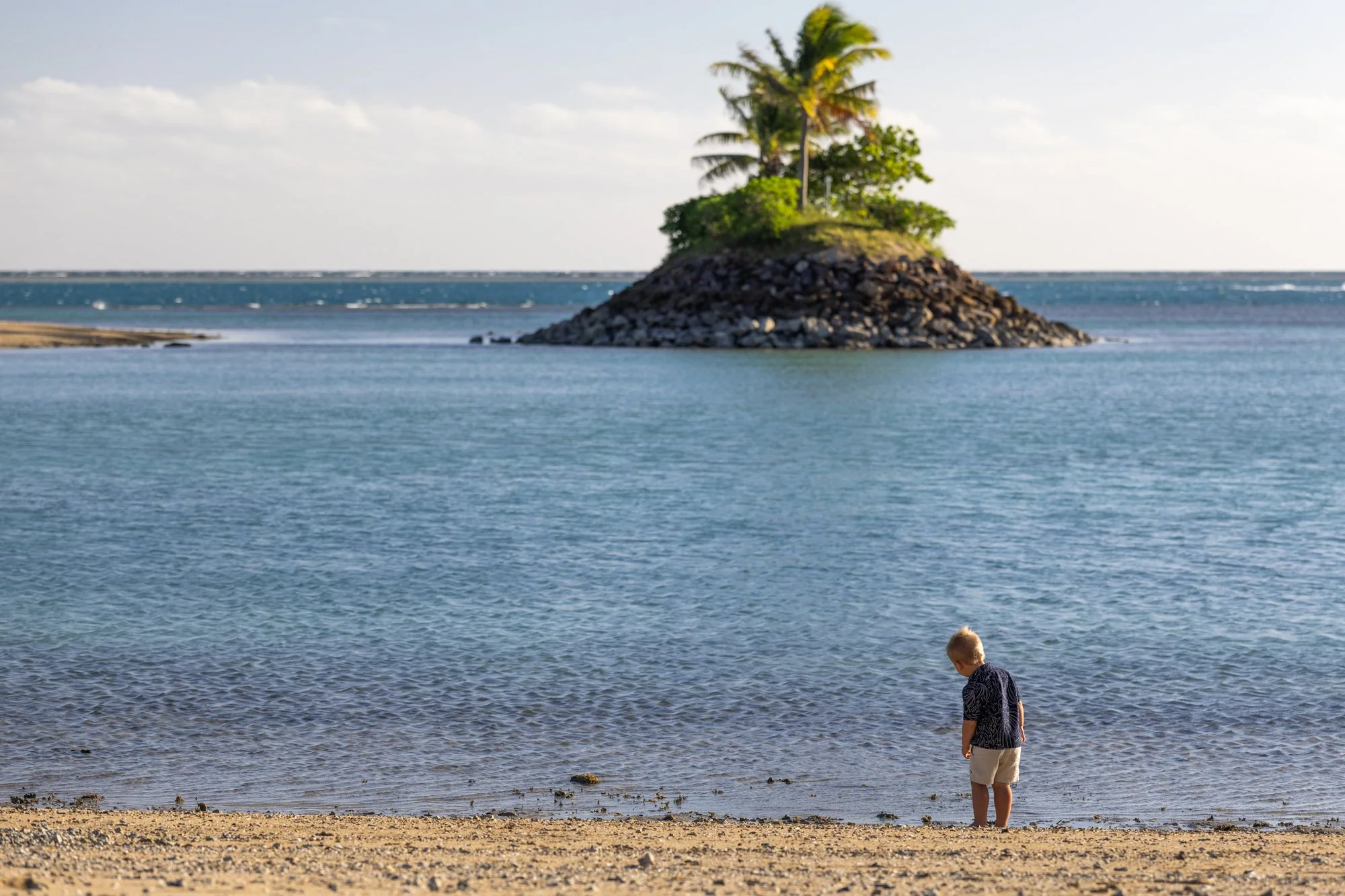 A young child explores the Fiji shoreline beneath a tiny palm-covered island.