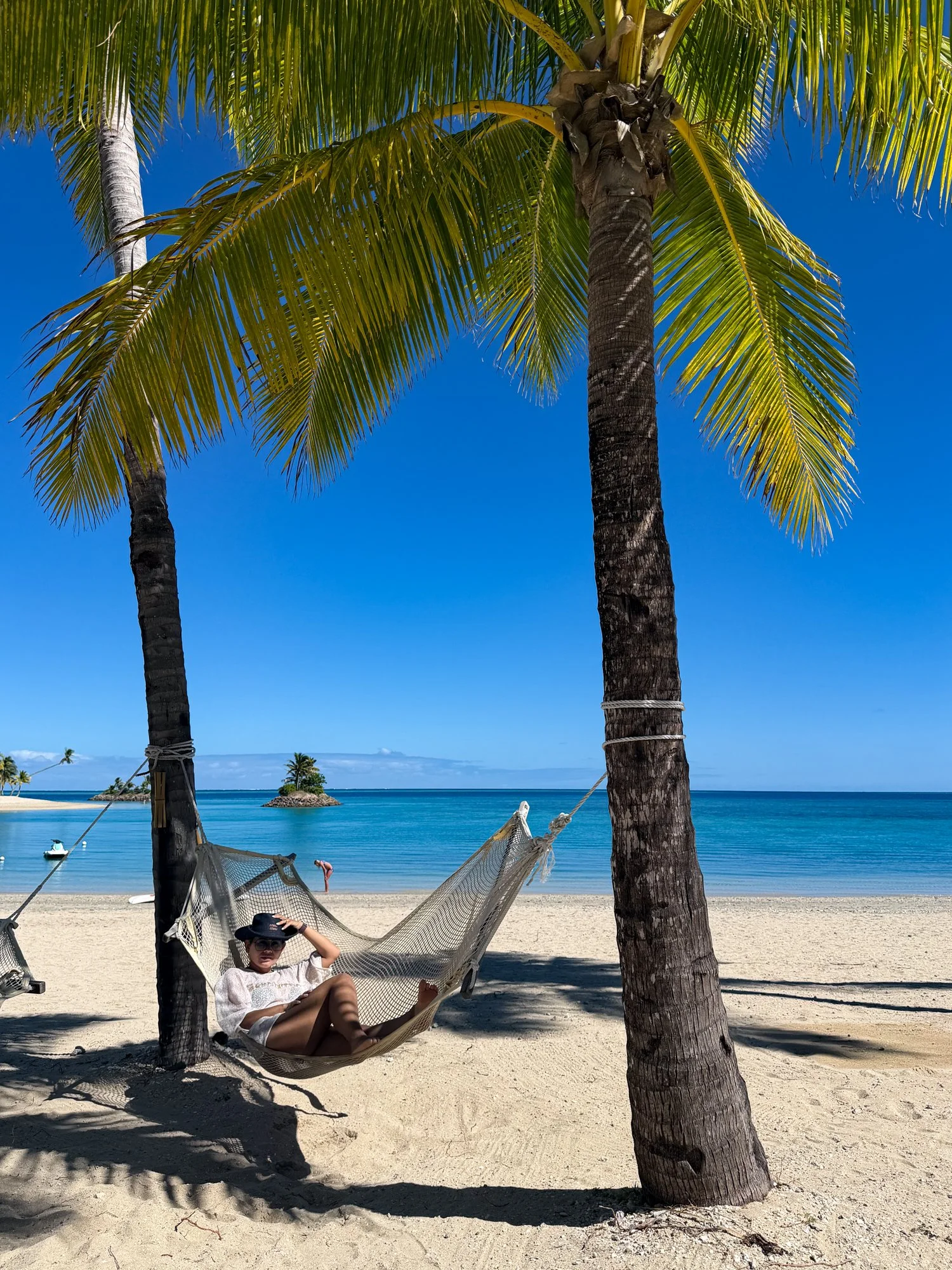 A traveler relaxes in a hammock beneath palm trees beside a turquoise Fiji beach.