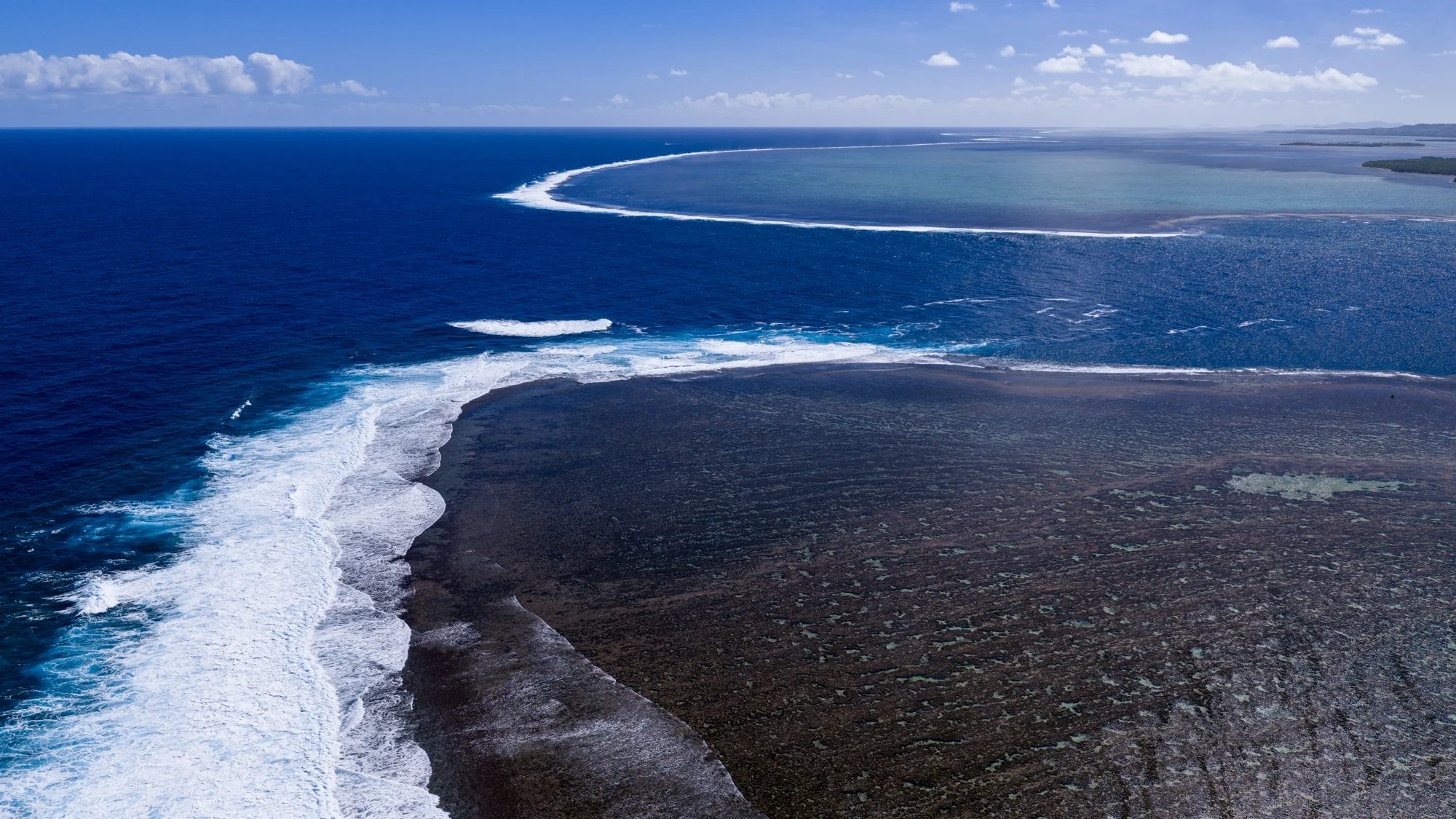 Aerial view of white waves curling around a vast offshore coral reef in Fiji.