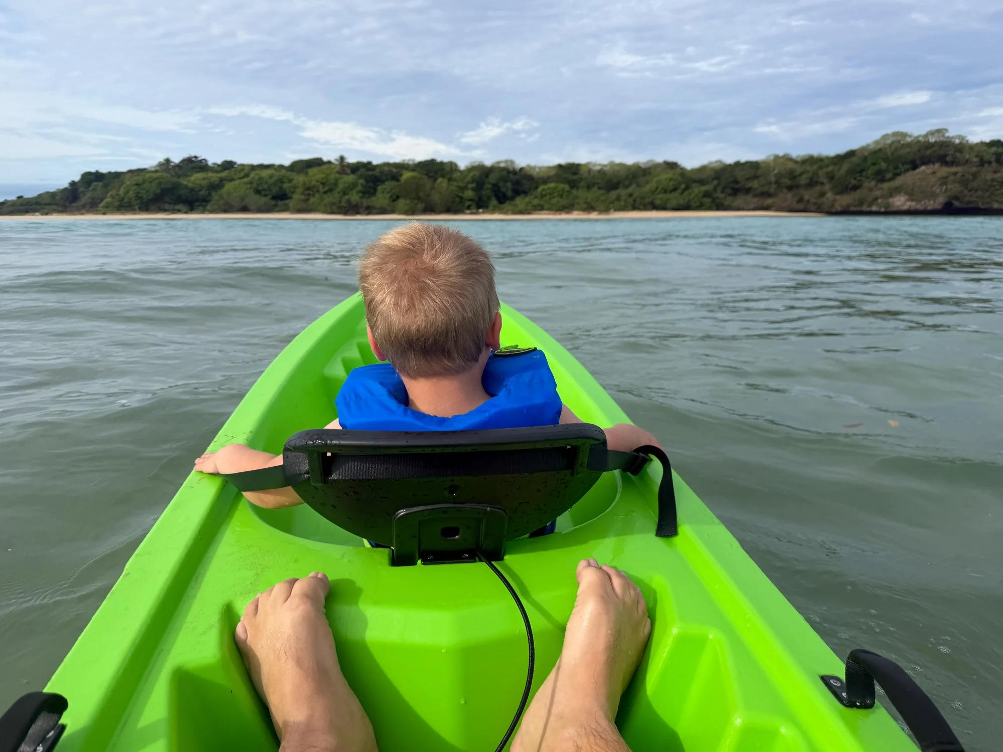 A young child rides in the front of a bright green kayak toward a forested Fiji shoreline.