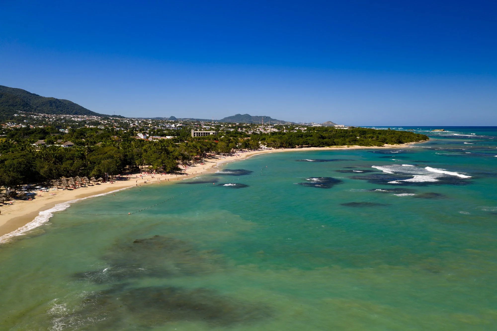 Aerial view of a turquoise beach, palm-lined coast, and green mountains near Puerto Plata, Dominican Republic.