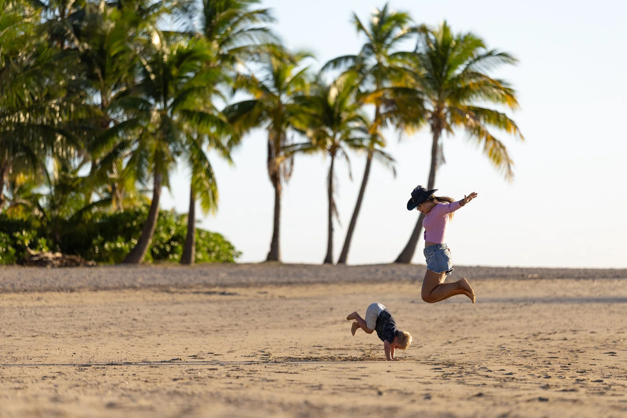 A traveler and young child leap and tumble on a palm-lined Fiji beach at golden hour.