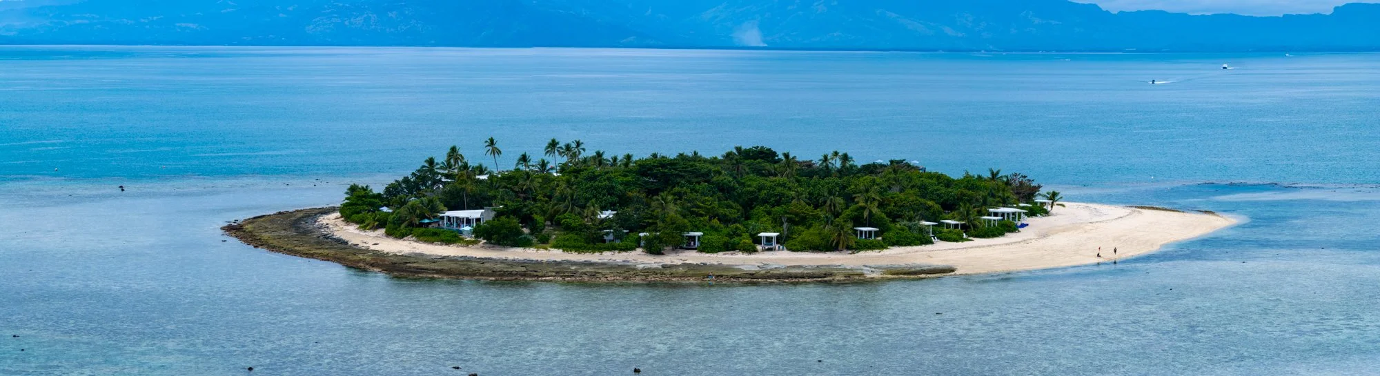 Panoramic aerial view of a small Fijian island resort with a white-sand spit and distant mountains.