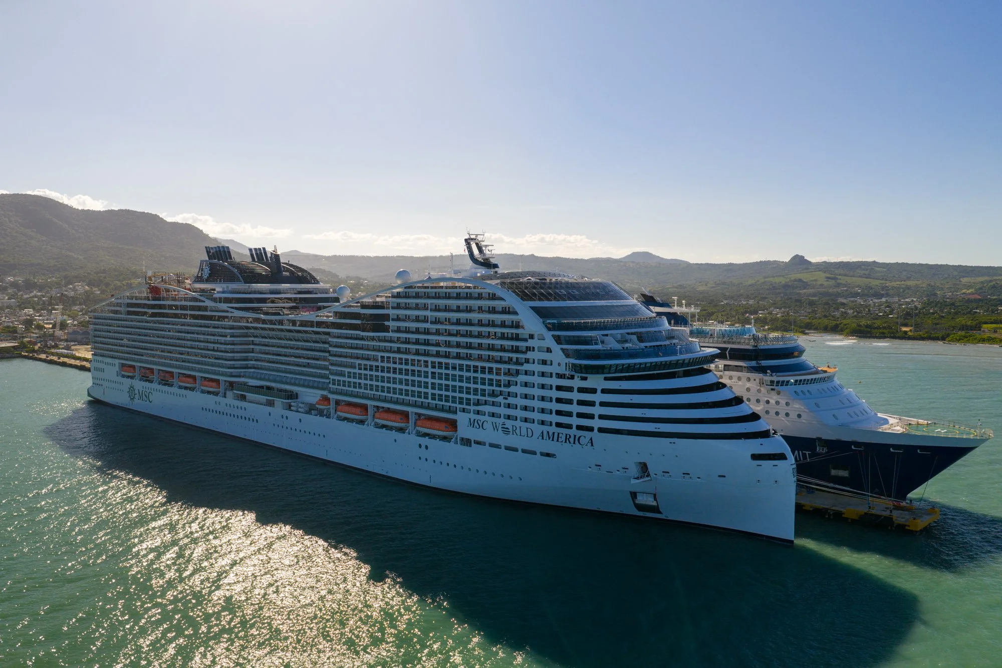 MSC World America docked beside another cruise ship at Puerto Plata with mountains in the background.
