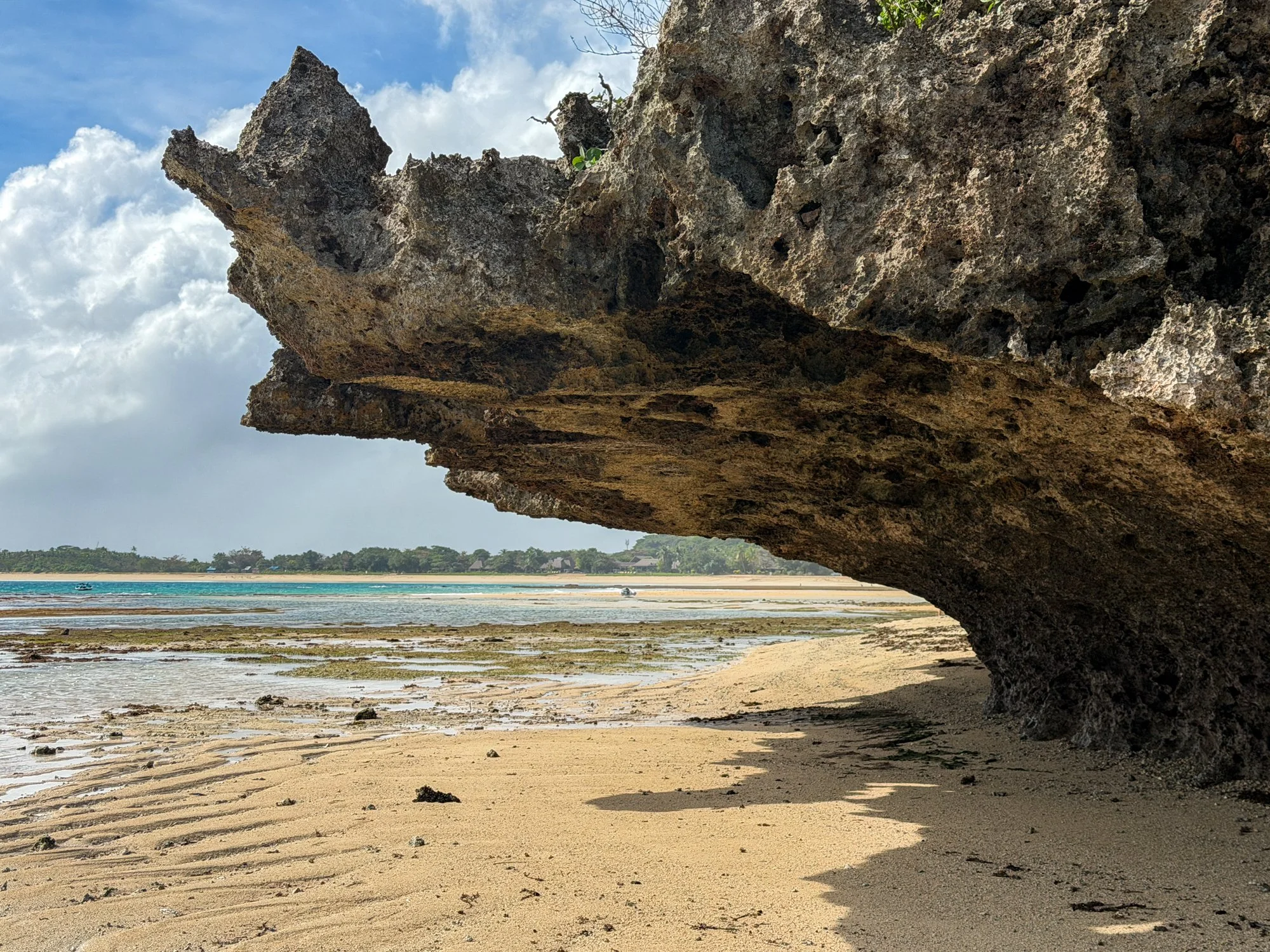 A dramatic limestone overhang casts a deep shadow across a Fiji beach at low tide.