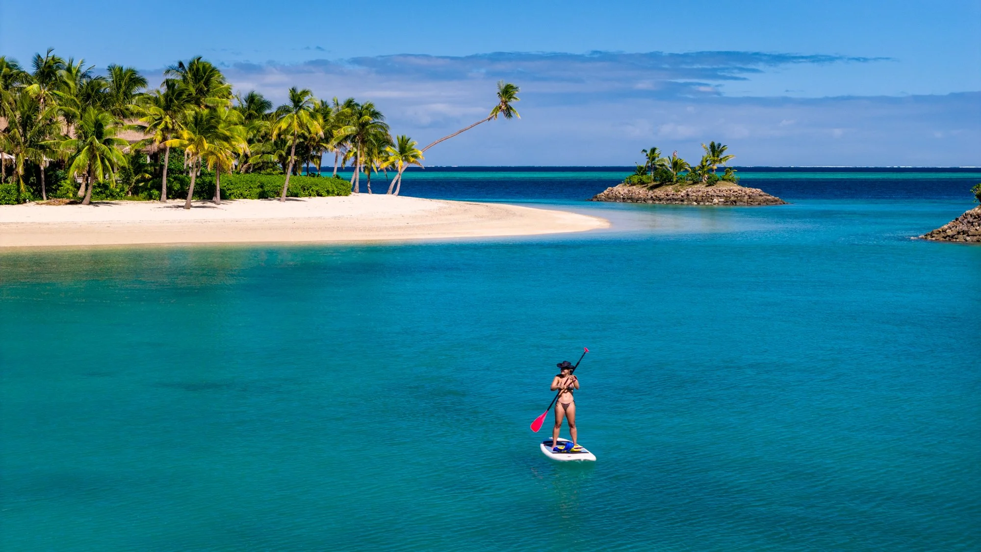 A paddleboarder glides across a calm turquoise lagoon beside a palm-lined beach in Fiji.