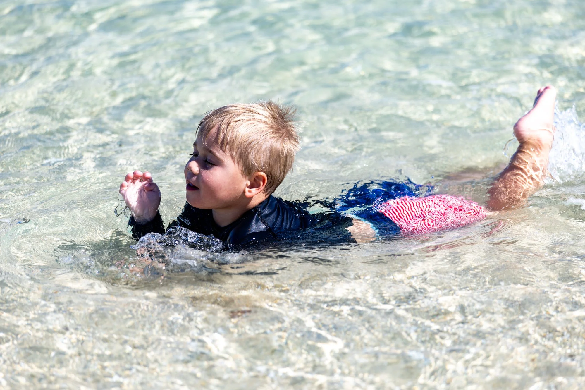 A young child swims through sunlit, crystal-clear shallows on a Fiji beach.