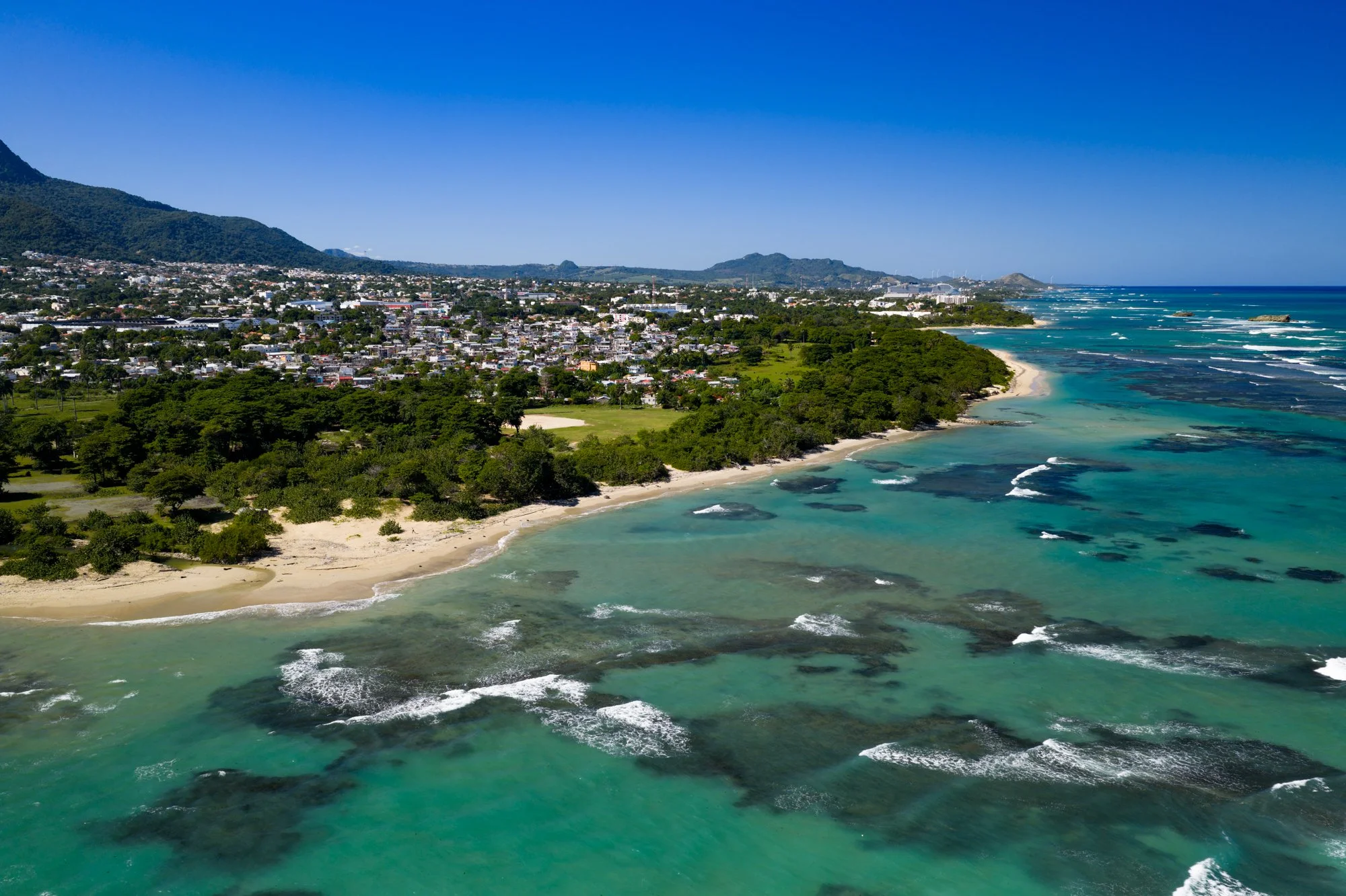 Aerial view of Puerto Plata’s coastline, coral shallows, and neighborhoods beneath green mountains.