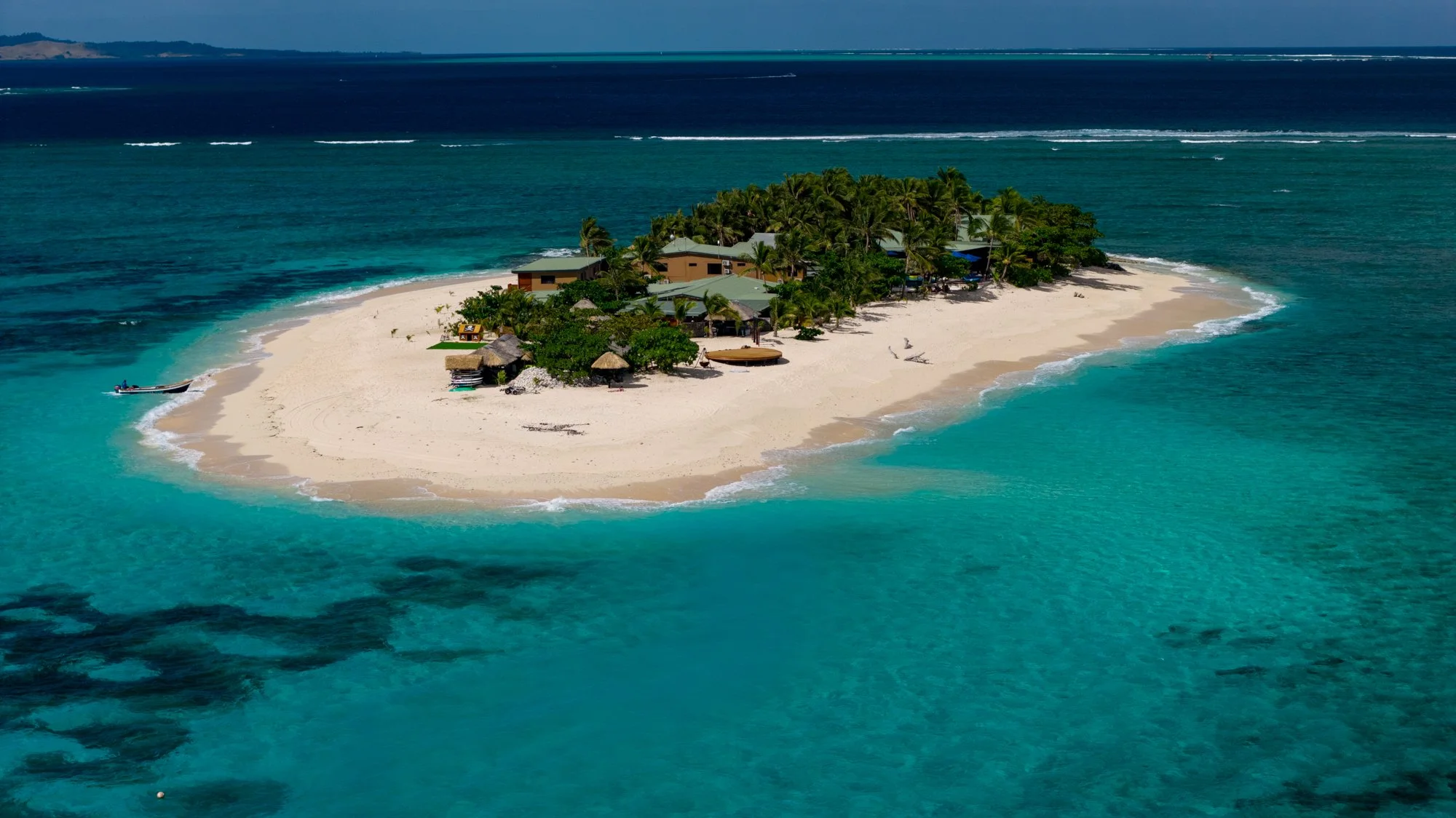 Aerial view of a tiny palm-covered island ringed by white sand and clear turquoise water in Fiji.