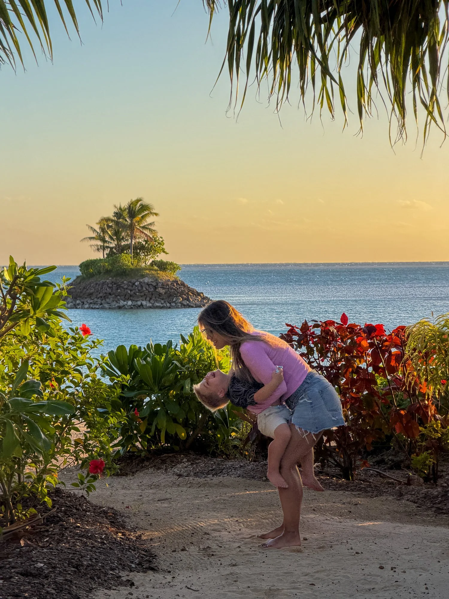 A traveler playfully dips a laughing young child in a tropical garden at golden hour in Fiji.