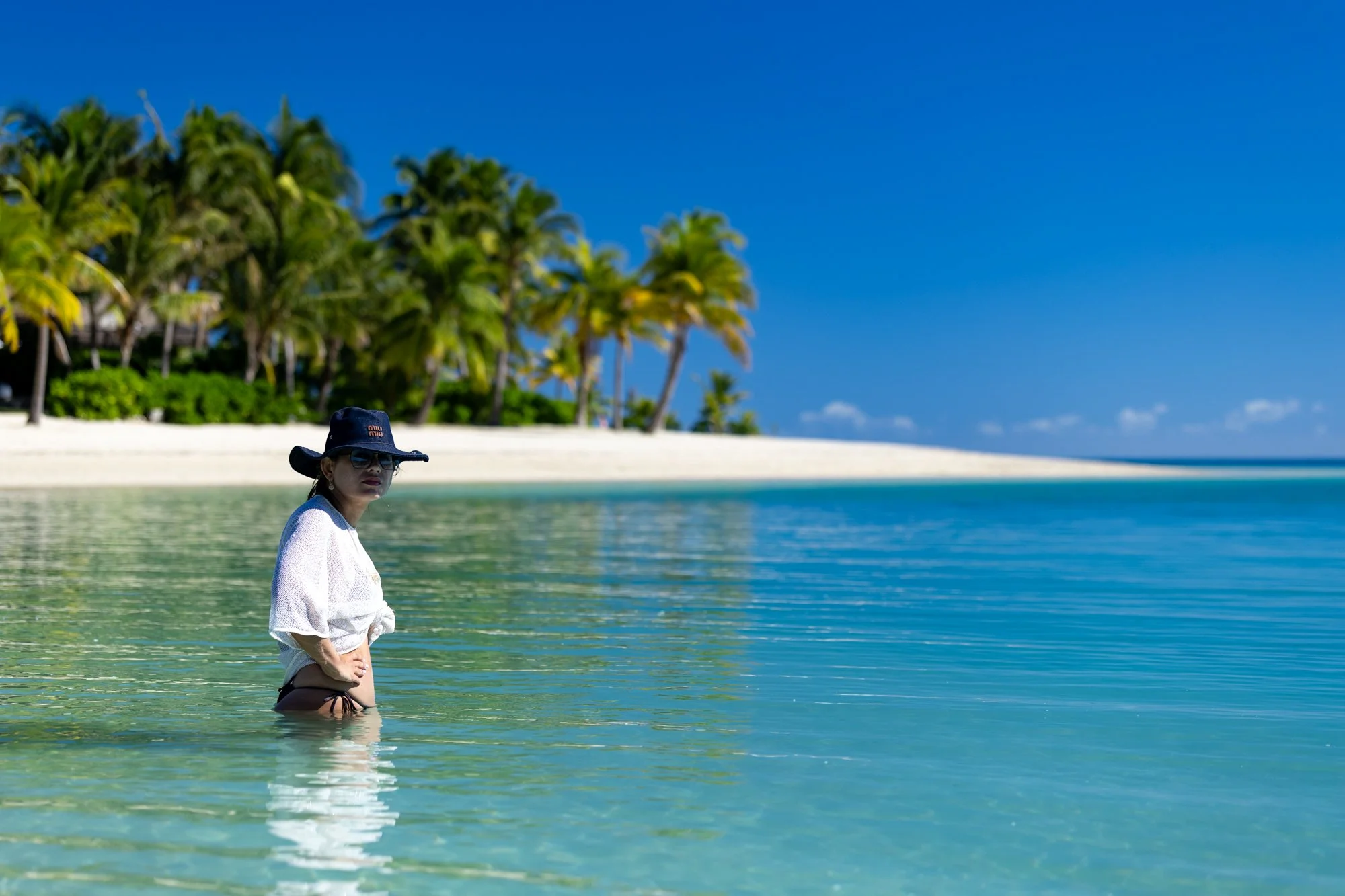A traveler in a sunhat stands waist-deep in the calm turquoise water of a Fijian lagoon.