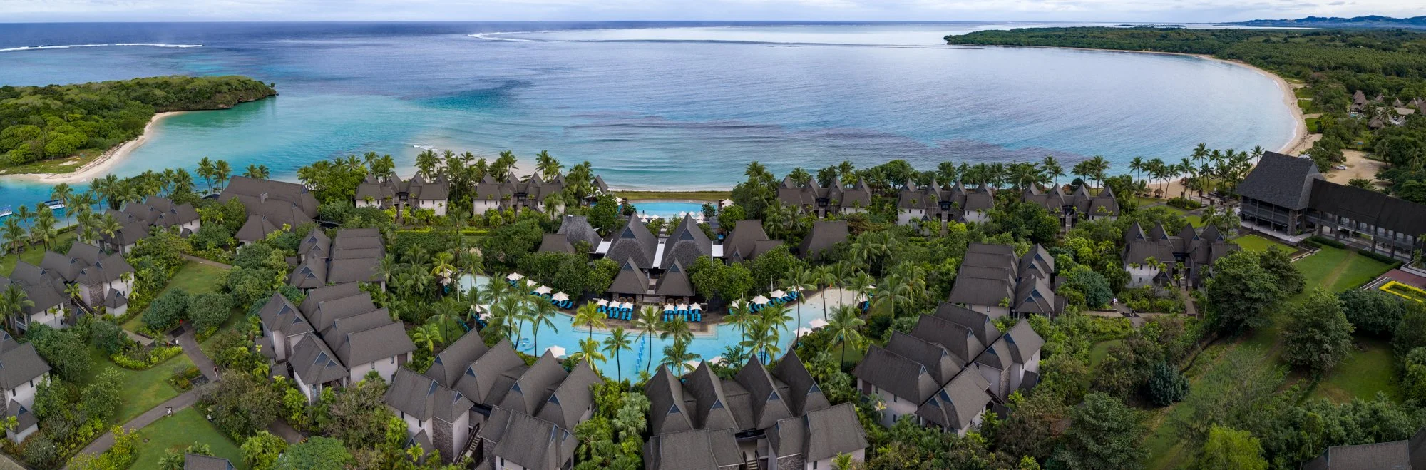 Aerial panorama of a beachfront Fiji resort with tropical gardens, pools, and a calm bay.