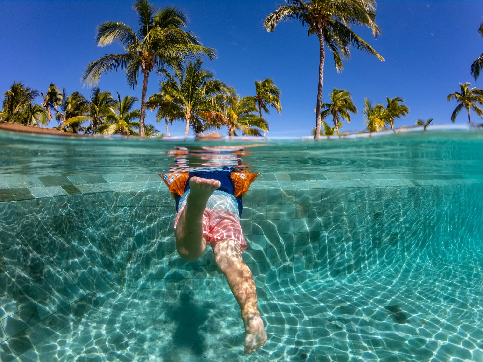 Split-level view of a young child swimming in a tropical resort pool beneath Fiji palm trees.