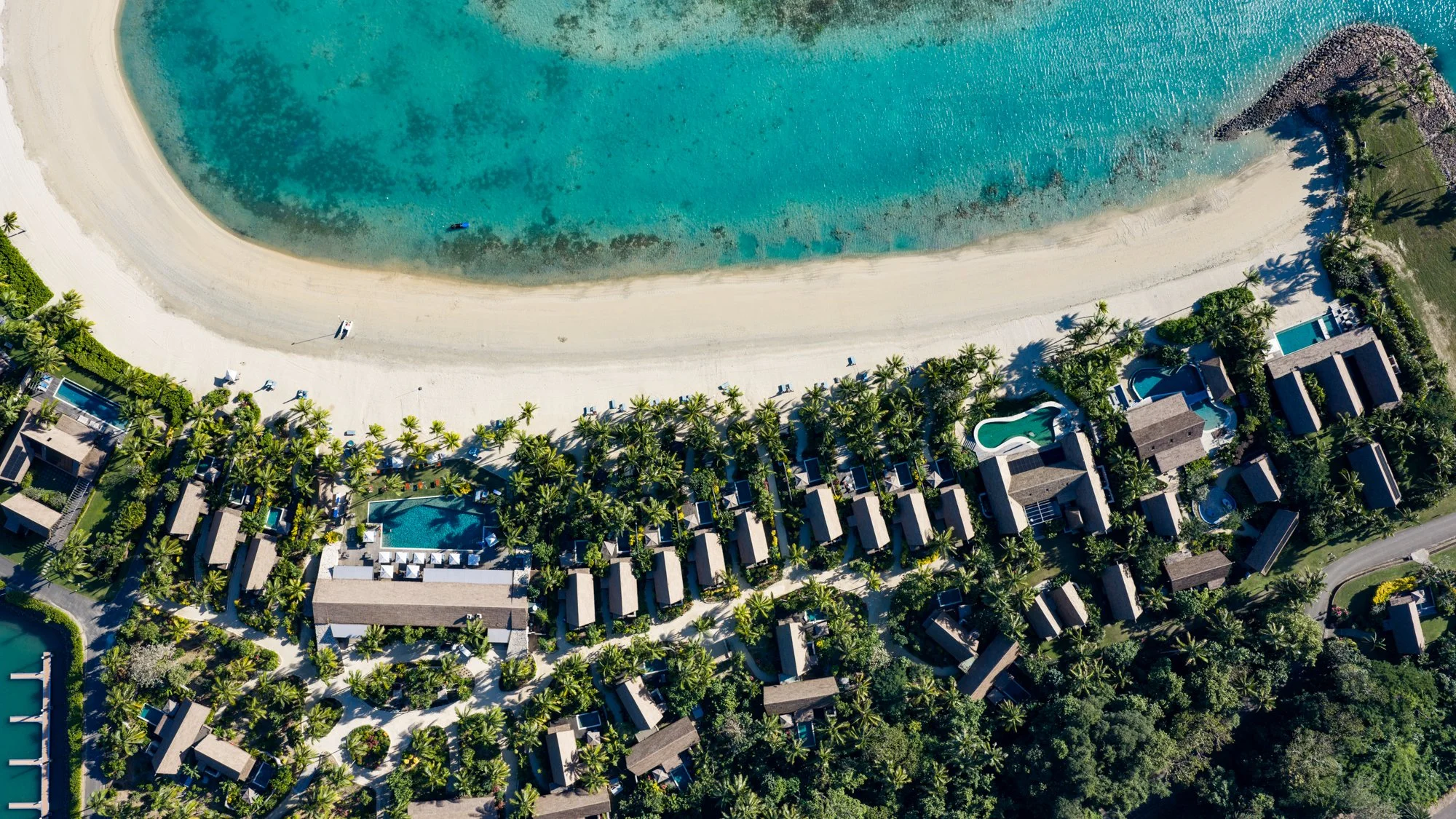 Top-down aerial view of a palm-filled beachfront resort, pools, and a curving turquoise lagoon in Fiji.