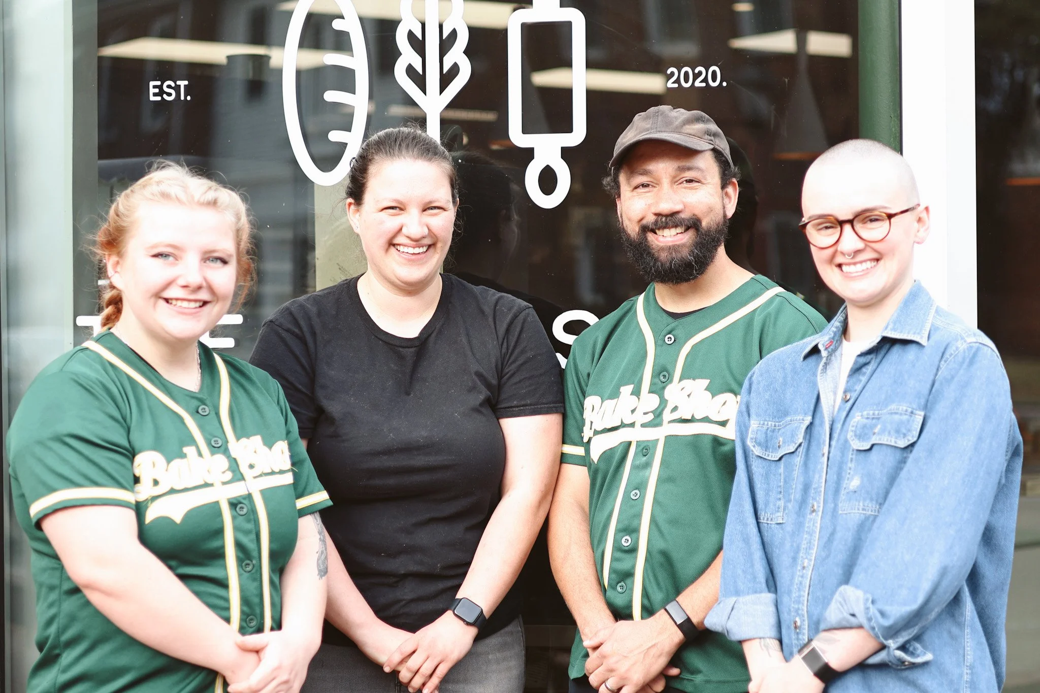 Four smiling people standing in front of a glass door with decorative white graphics; two women dressed in green baseball jerseys, one woman in a black t-shirt, and a man in a green baseball jersey with a cap.