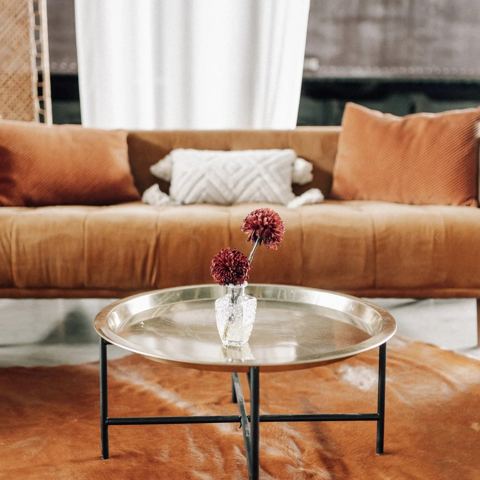 Modern living room with a round metal tray table holding a small glass vase with three red flowers, in front of a tan leather sofa with orange and white pillows, and a cowhide rug on the floor.