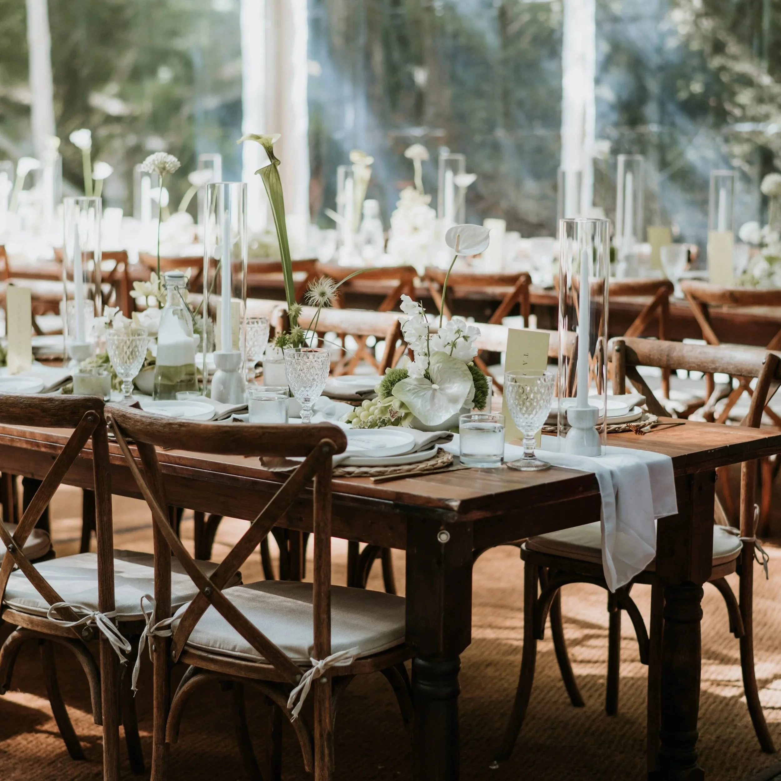 A beautifully decorated outdoor dining table with white floral centerpieces, tall glass candle holders, and place settings with white plates and napkins, set in a wooded area with natural light.
