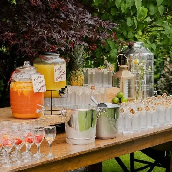 Sweet tea, lemonade, and peach juice dispensers on a wooden table with glassware, pineapples, limes, a lantern, and decorative jars, set outdoors with green foliage in the background.