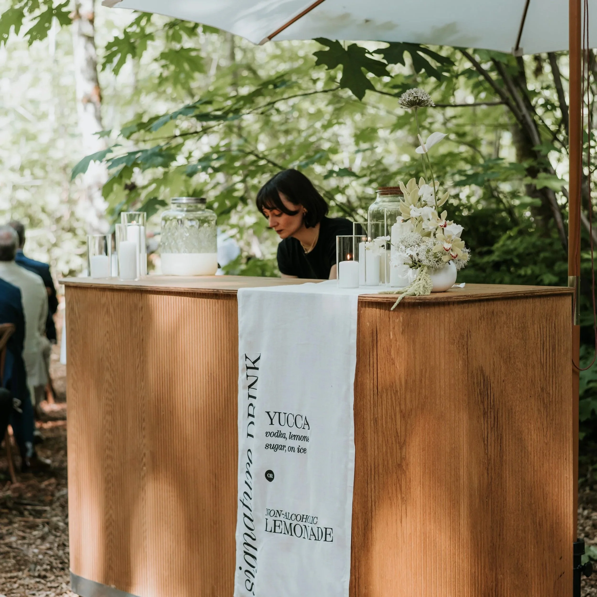 A woman standing behind a wooden beverage stand decorated with white flowers and candles under a large umbrella, with lush green trees in the background.