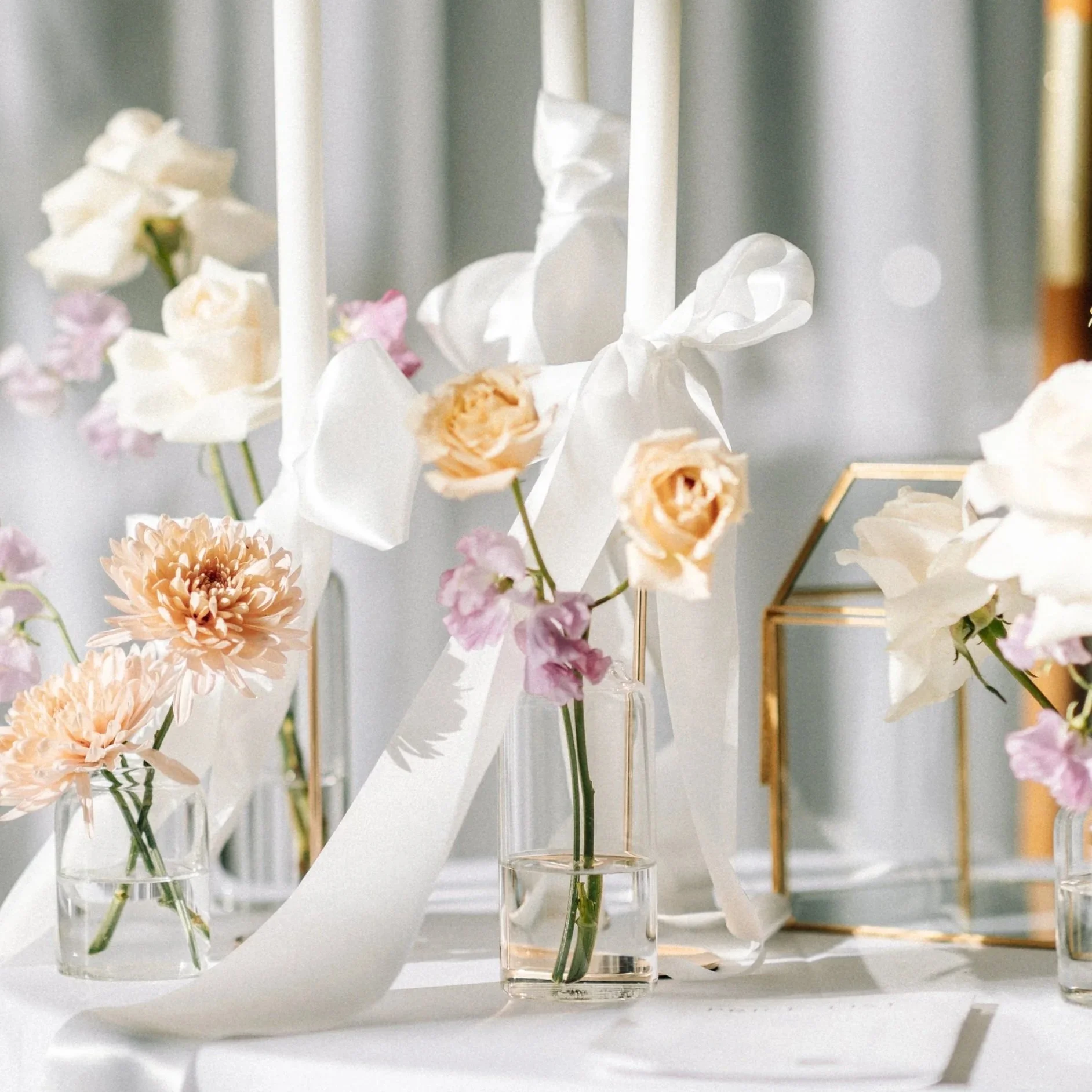 Arrangement of pastel-colored flowers in glass vases, decorative white ribbons, and a geometric glass box on a white surface with soft natural lighting.