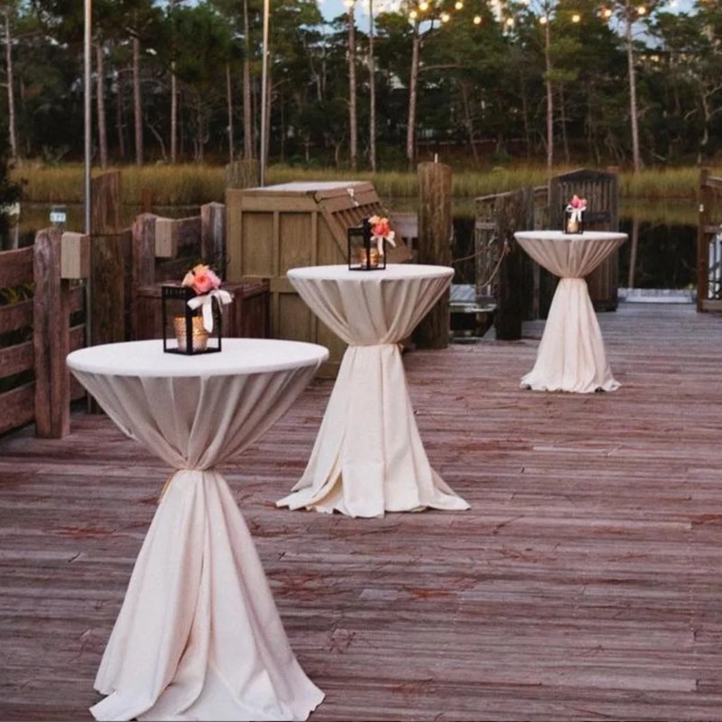 Three high-top tables with white tablecloths, decorated with small vases of pink flowers and lanterns, set on a wooden deck near a water body with trees in the background.