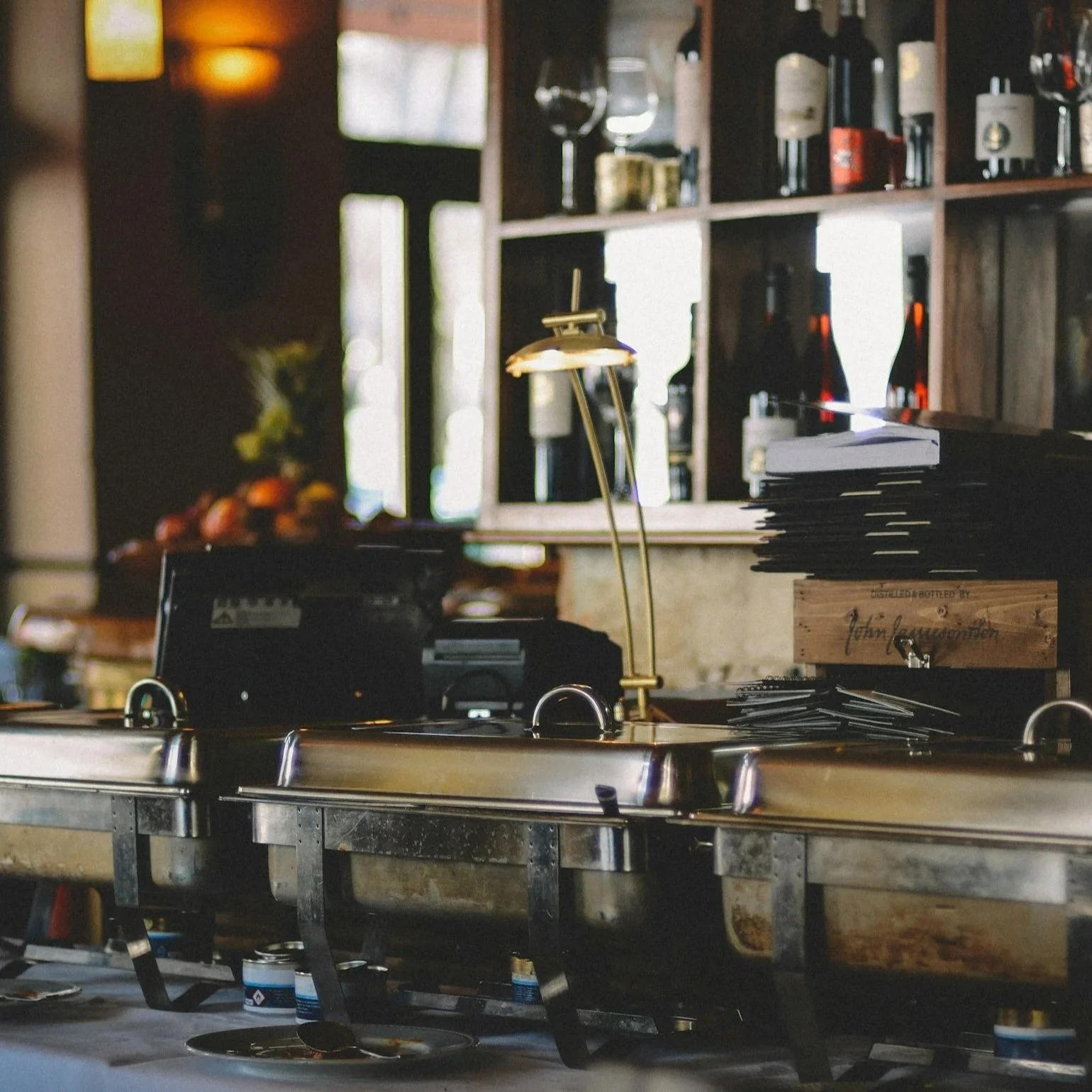 A buffet table with chafing dishes in a restaurant or cafe, with shelves of wine bottles and glasses in the background, and a stack of napkins or menus on the right side.