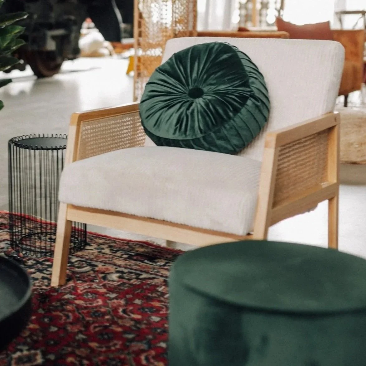 A modern chair with a white fabric cushion and wooden frame, decorated with a green velvet round pillow, placed on a colorful patterned rug in a cozy interior setting.