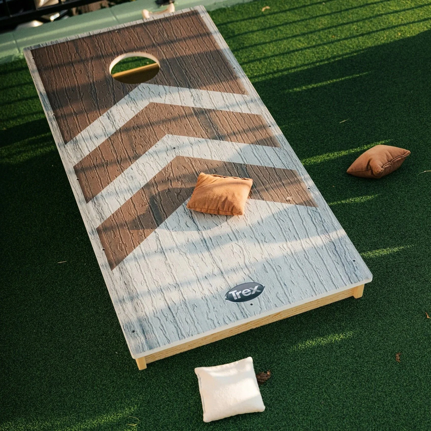 A cornhole game board with brown and white stripes on a green artificial turf surface, four bean bags around it, and a metal railing in the background.