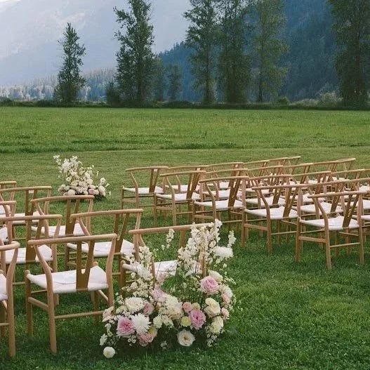 Empty outdoor wedding ceremony setup with wooden chairs, floral arrangements, and a lush green field with trees in the background.