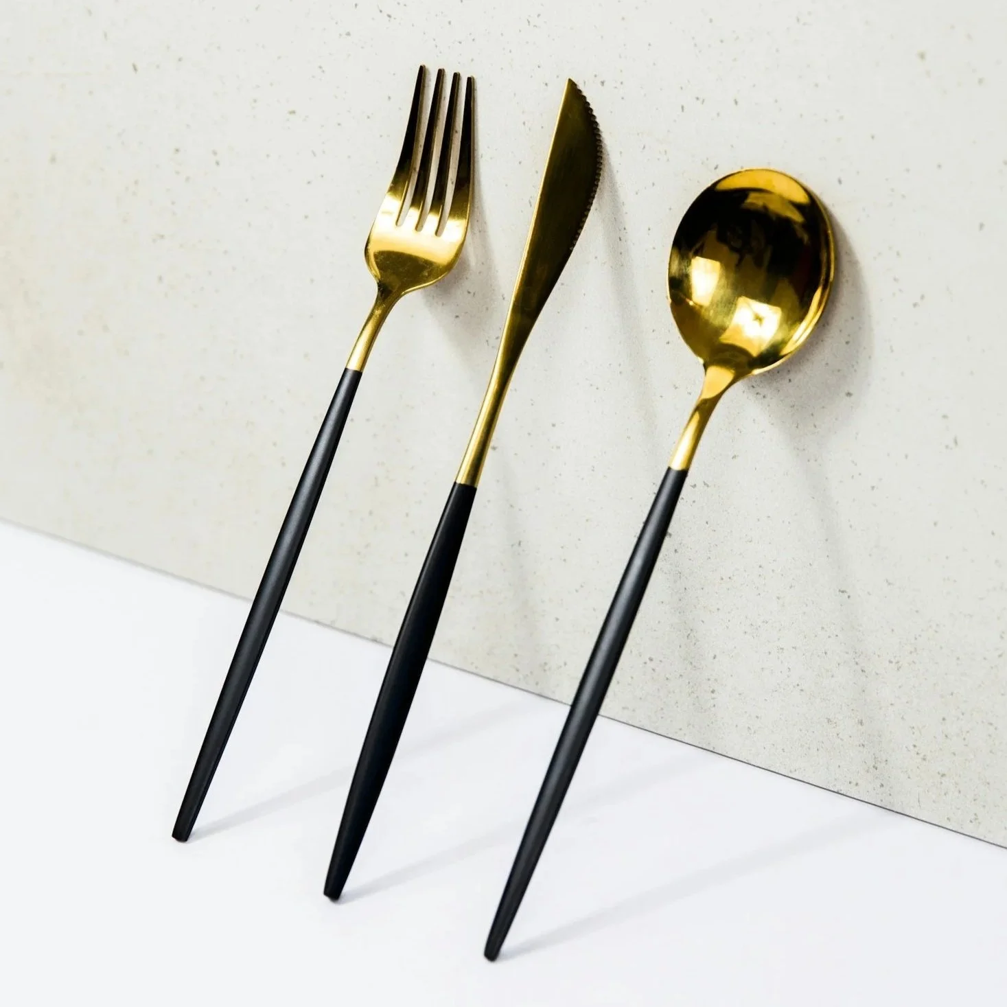 A set of modern gold and black cutlery, including a fork, a knife, and a spoon, arranged on a white surface and against a beige speckled background.