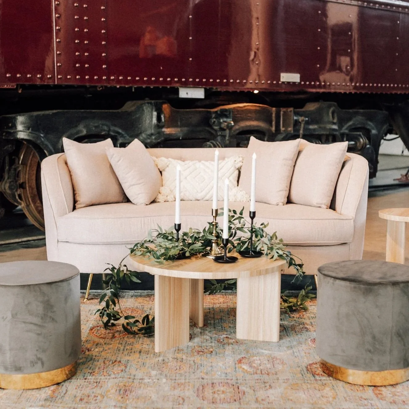 Living room setting with a beige sofa, four cushions, a small wooden coffee table with greenery and four white candles, two gray drum stools, a patterned area rug, and a dark train car in the background.