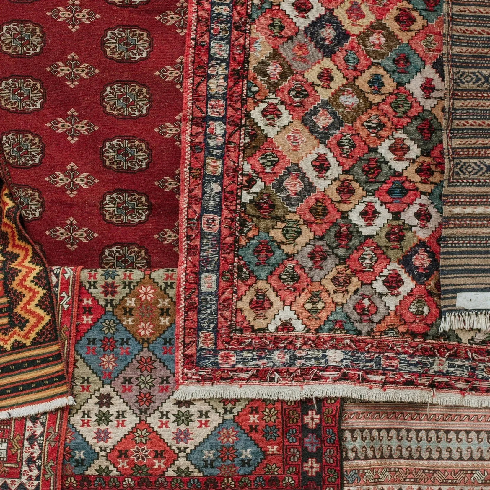 A collection of overlapping traditional Persian-style rugs with intricate geometric and floral patterns in red, blue, beige, and black colors.