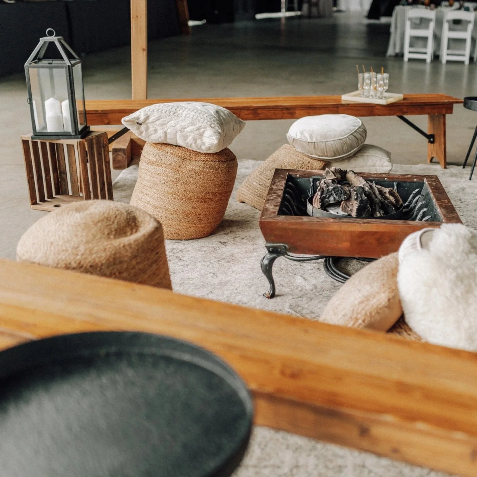 Cozy indoor seating area with woven poufs, pillows, a wooden bench, and a fire pit table with logs, decorated with candles and lanterns.