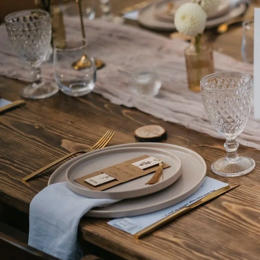 Elegant place setting with a wooden table, glass goblets, a white dinner plate, a beige napkin, gold flatware, and decorative tags with a tassel, with a centerpiece of white flowers in a small vase.