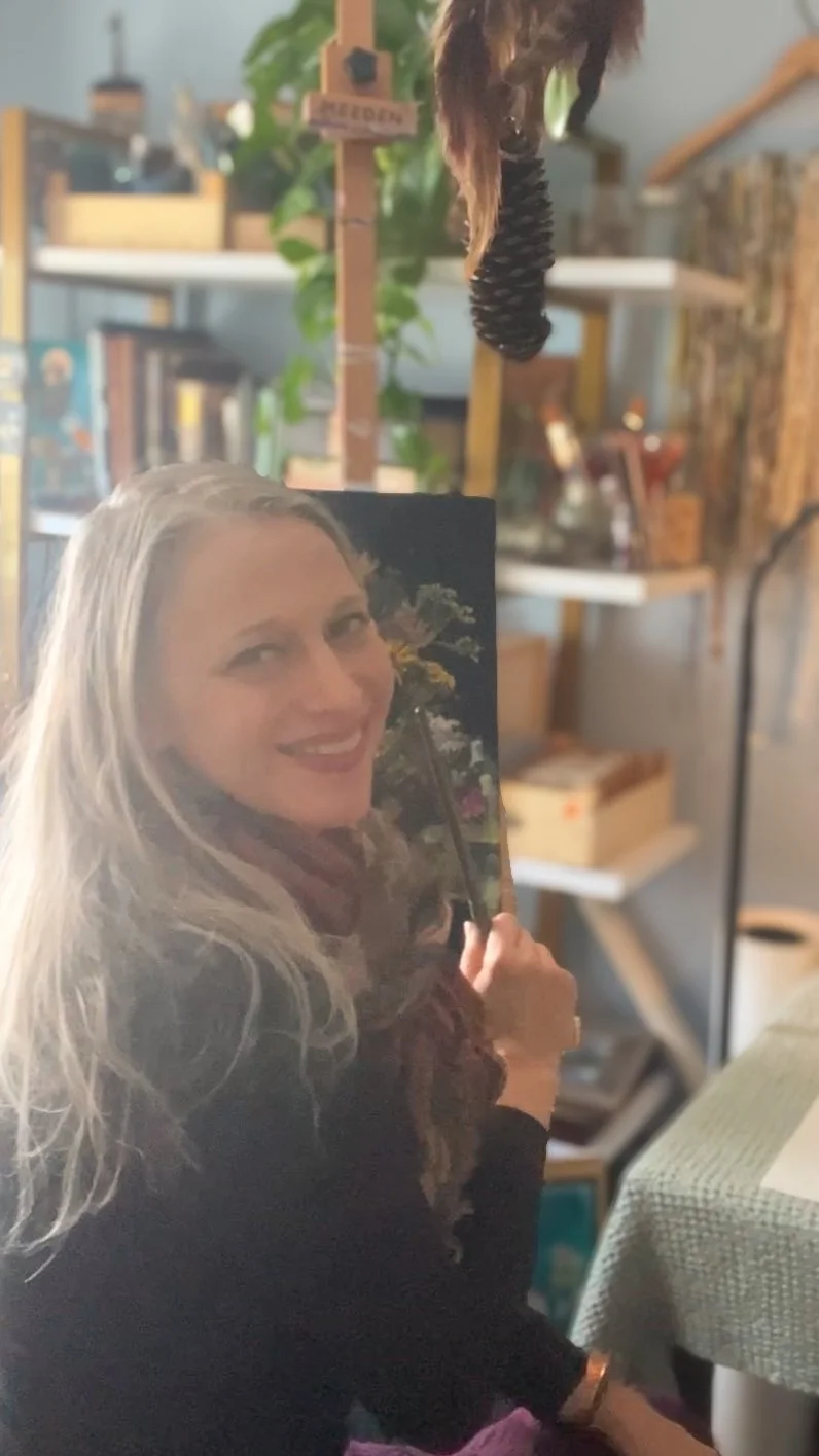 A woman with long blonde hair smiling while holding a mirror with a woman's face on it. The background shows shelves with plants and decorative items, creating a cozy, cluttered indoor space.