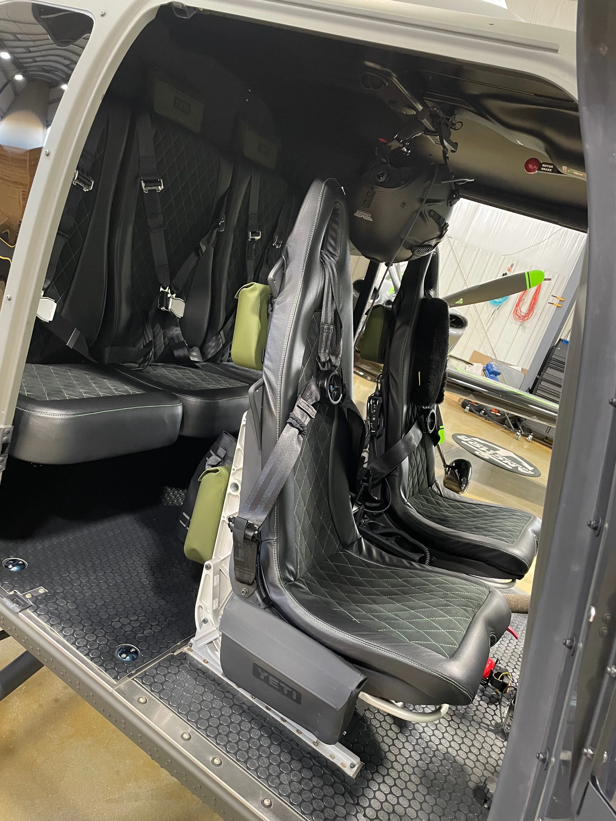 Interior of a small aircraft cockpit showing two black leather seats with diamond stitching, seat belts, and green pouches attached. The aircraft appears to be in a hangar with tools and equipment visible in the background.