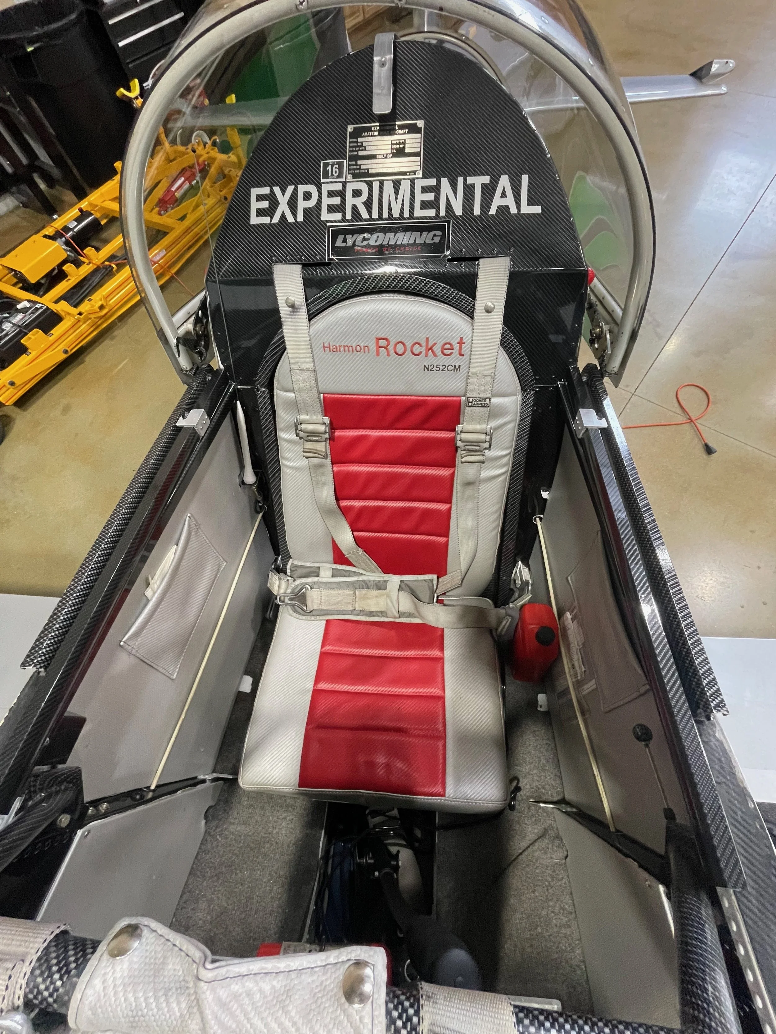 Interior view of a Harmon Rocket aircraft cockpit with a red and gray seat, safety harness, and various control panels and equipment.