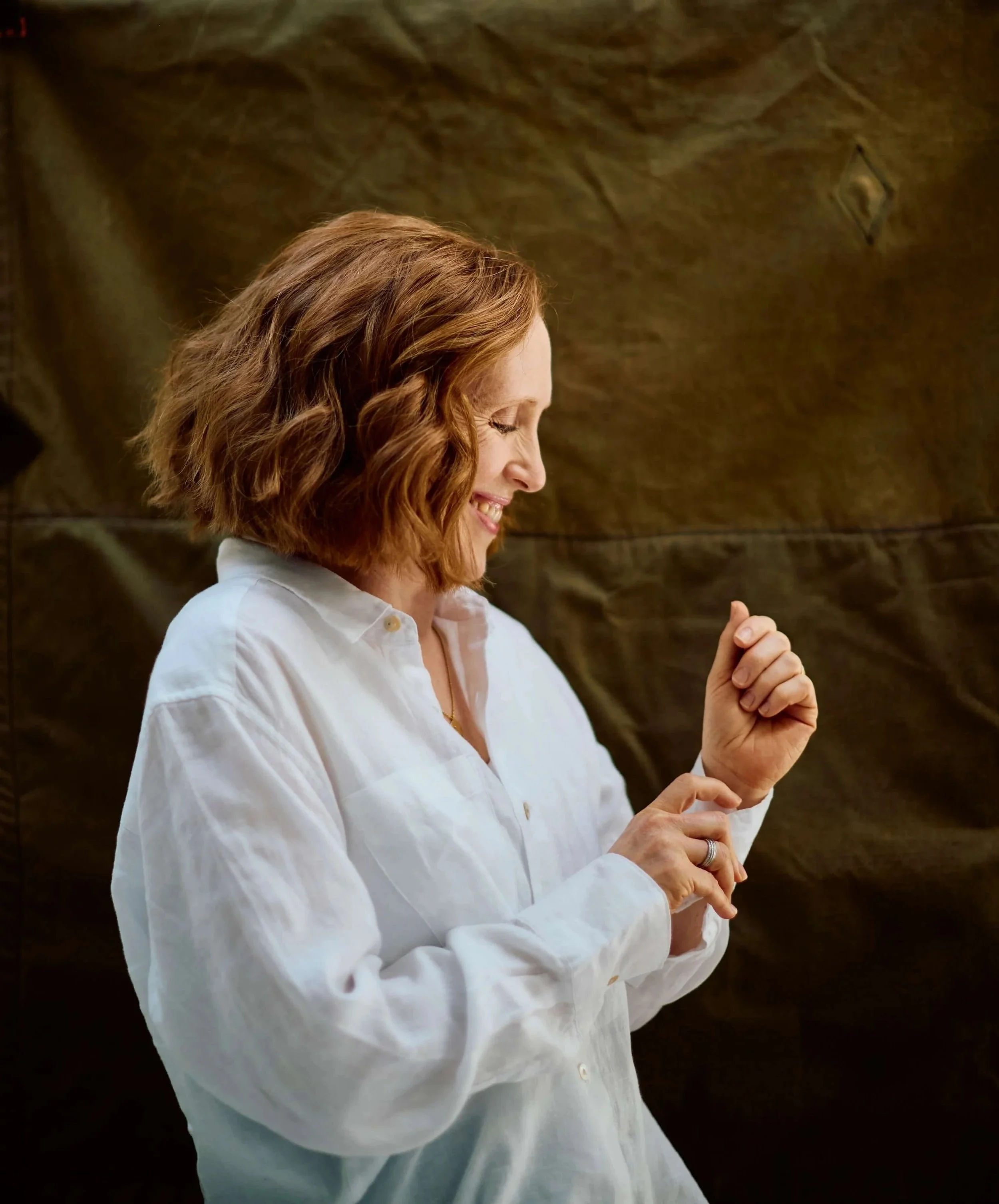 Woman with shoulder-length curly red hair smiling, wearing a white shirt, standing against a brown textured background.