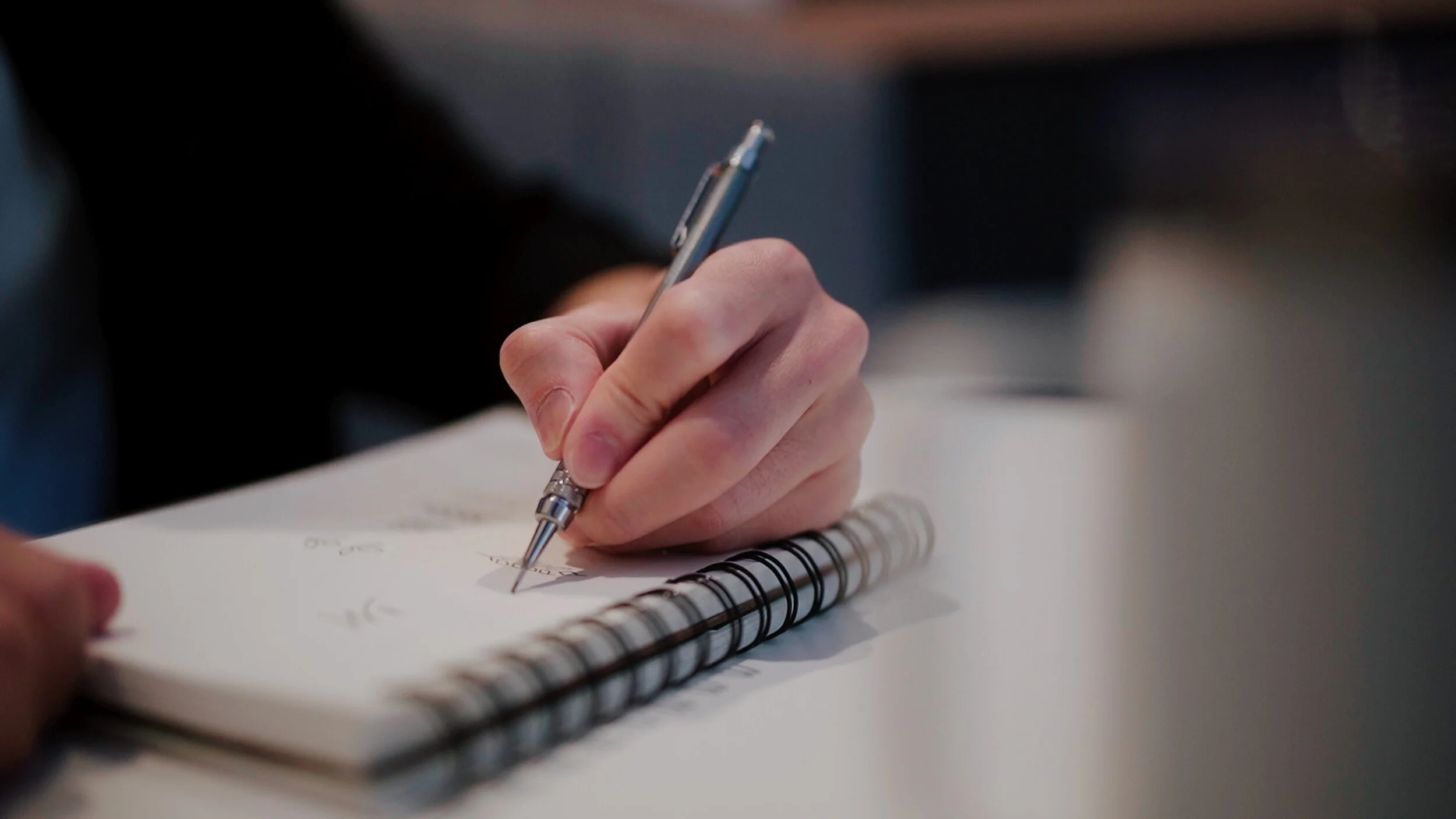 A person's hand holding a silver pen and writing in a spiral notebook on a white surface.